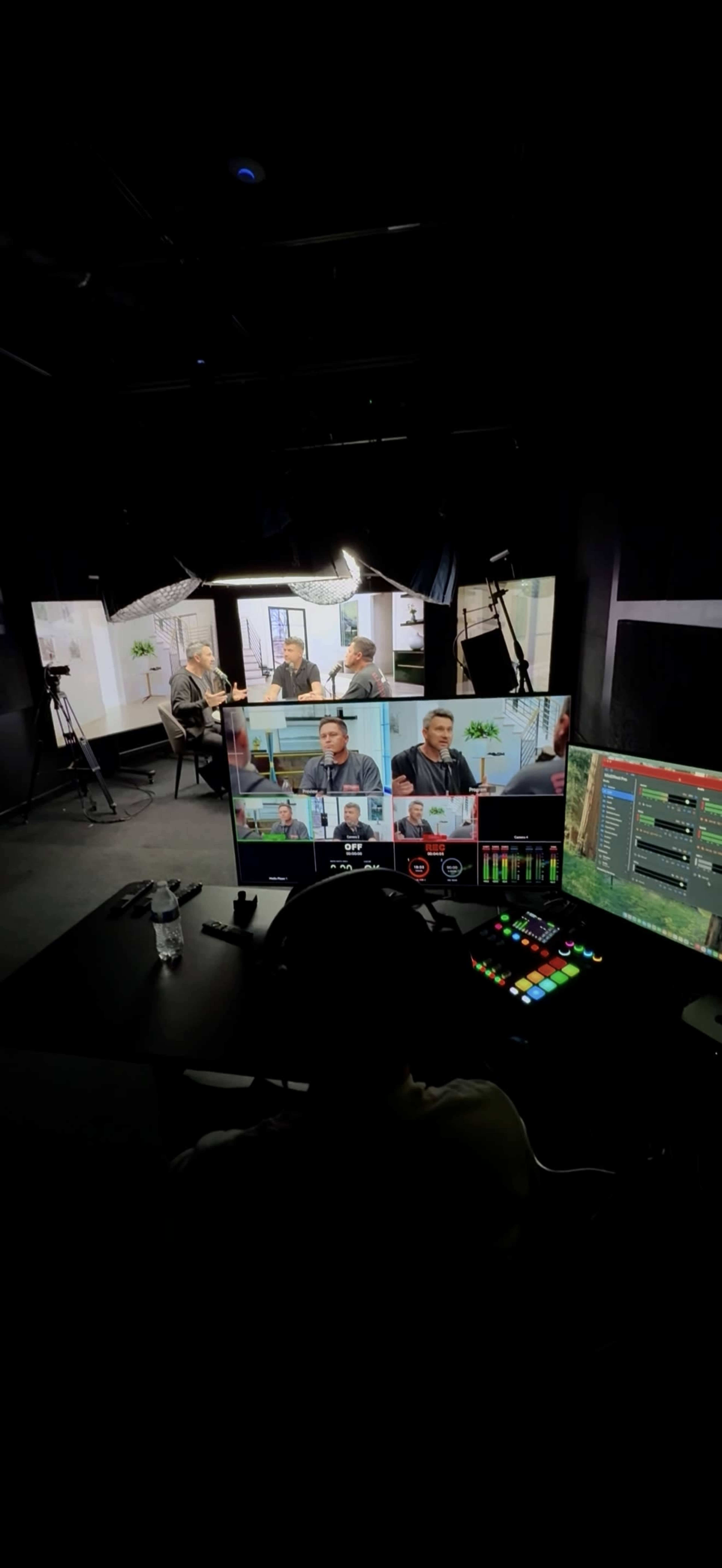 The image shows a studio setting with multiple screens displaying video feeds of a discussion among several individuals, while a person is operating equipment at a control desk.