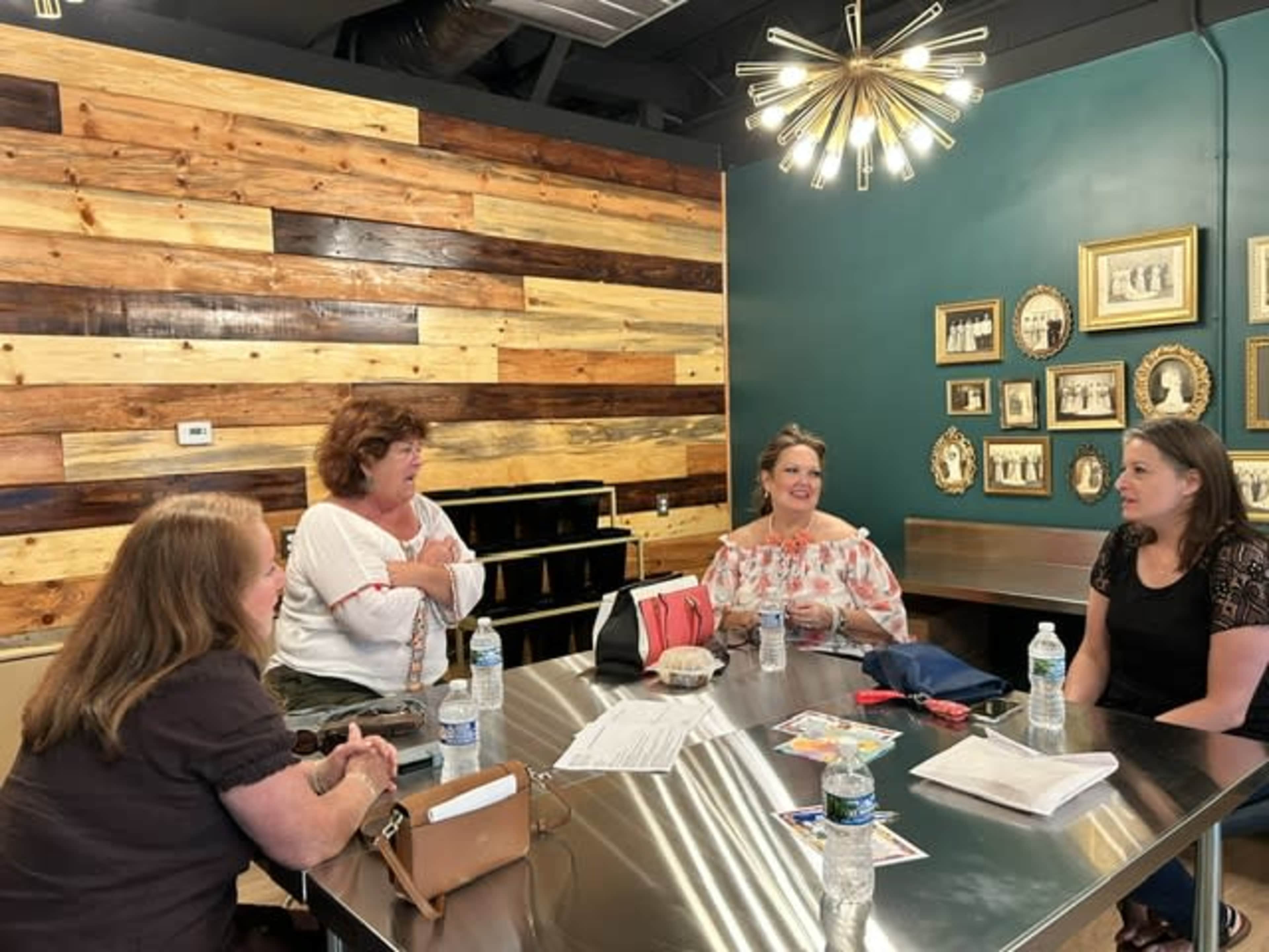 Four women sit around a metal table in a room with wooden walls and framed pictures, engaged in conversation.