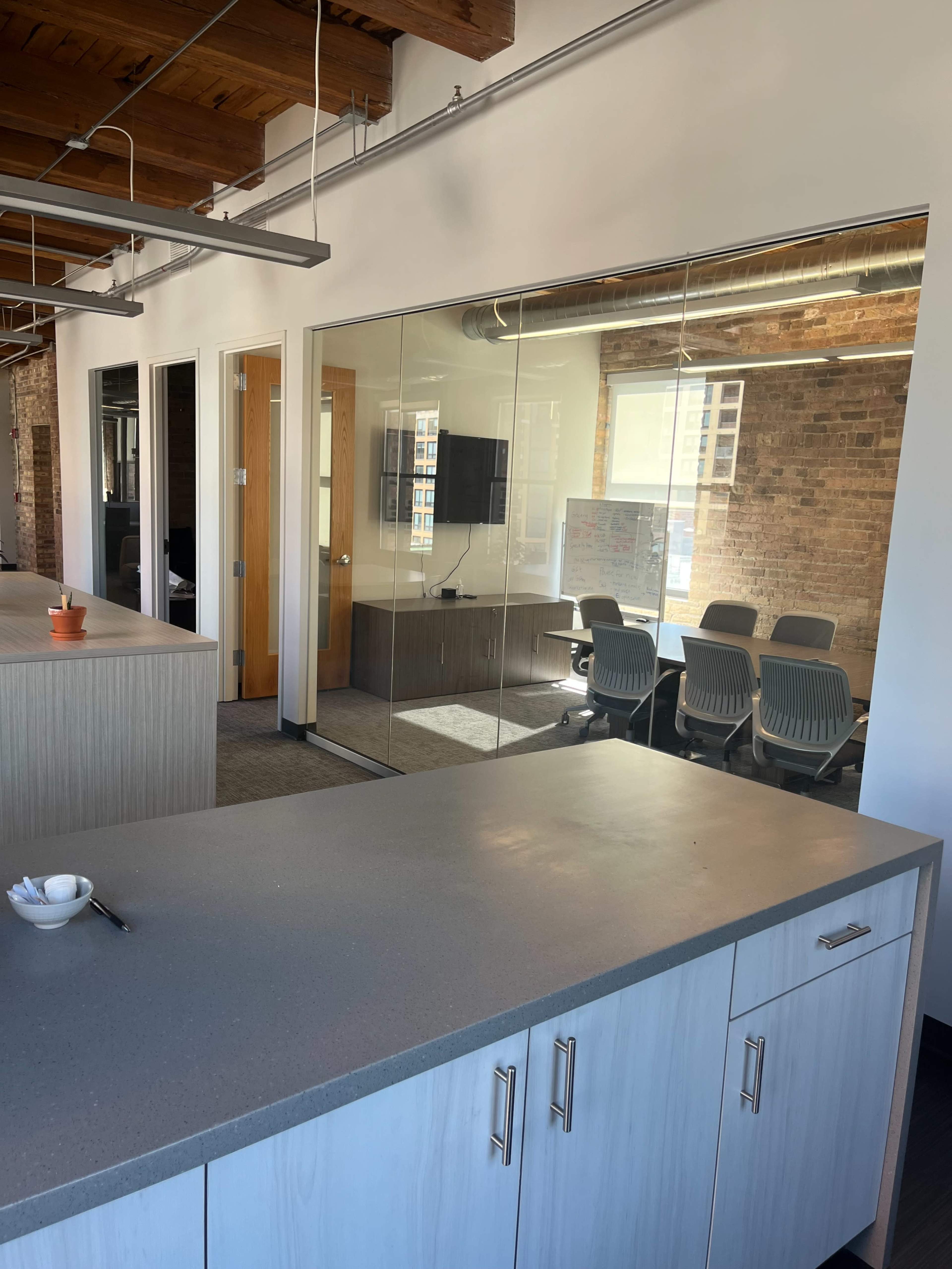 The image shows a modern office space with a gray countertop and a glass-walled conference room in the background.