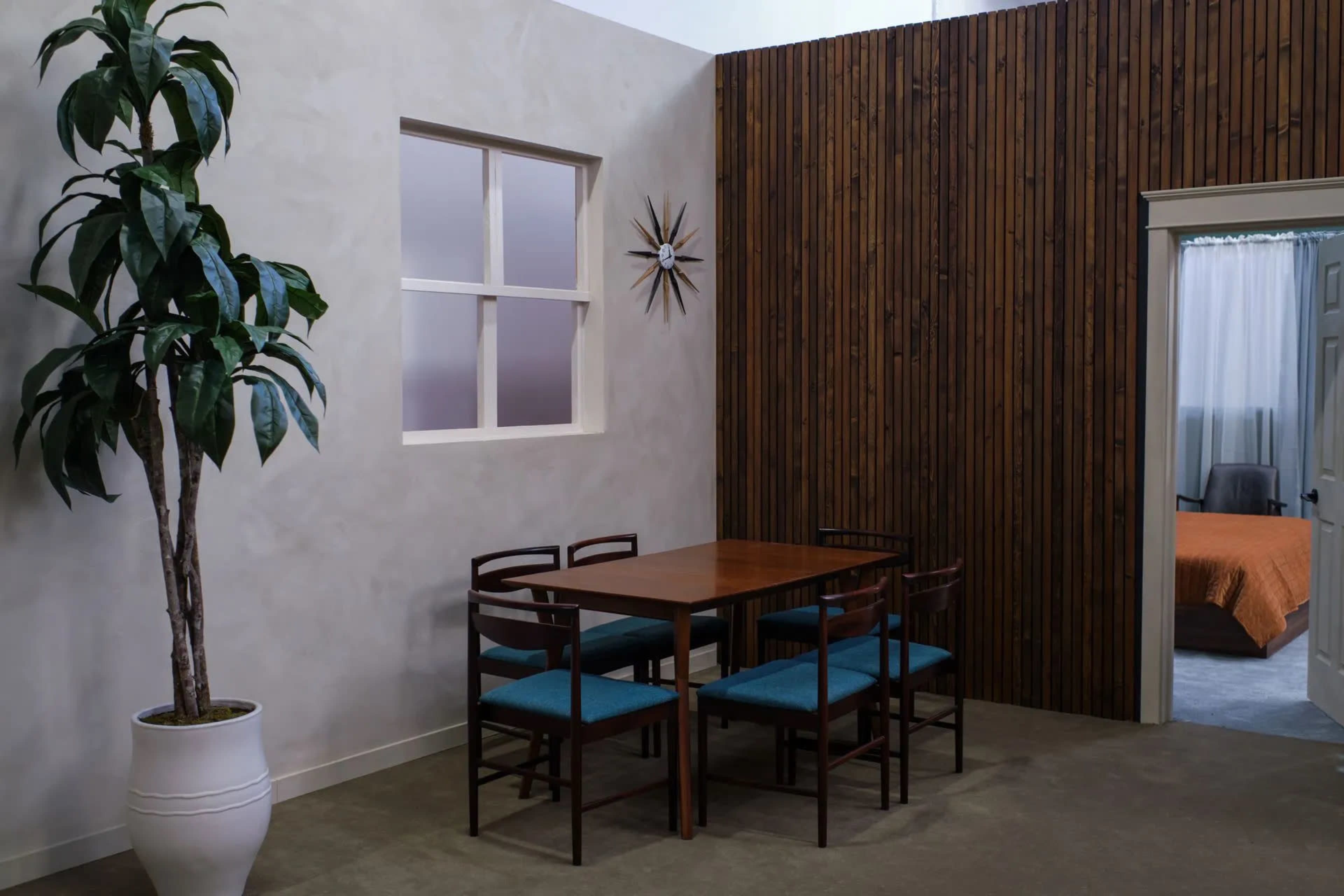 A dining area features a wooden table and four chairs, accompanied by a potted plant, with a wall made of wooden slats in the background and a doorway leading to another room.