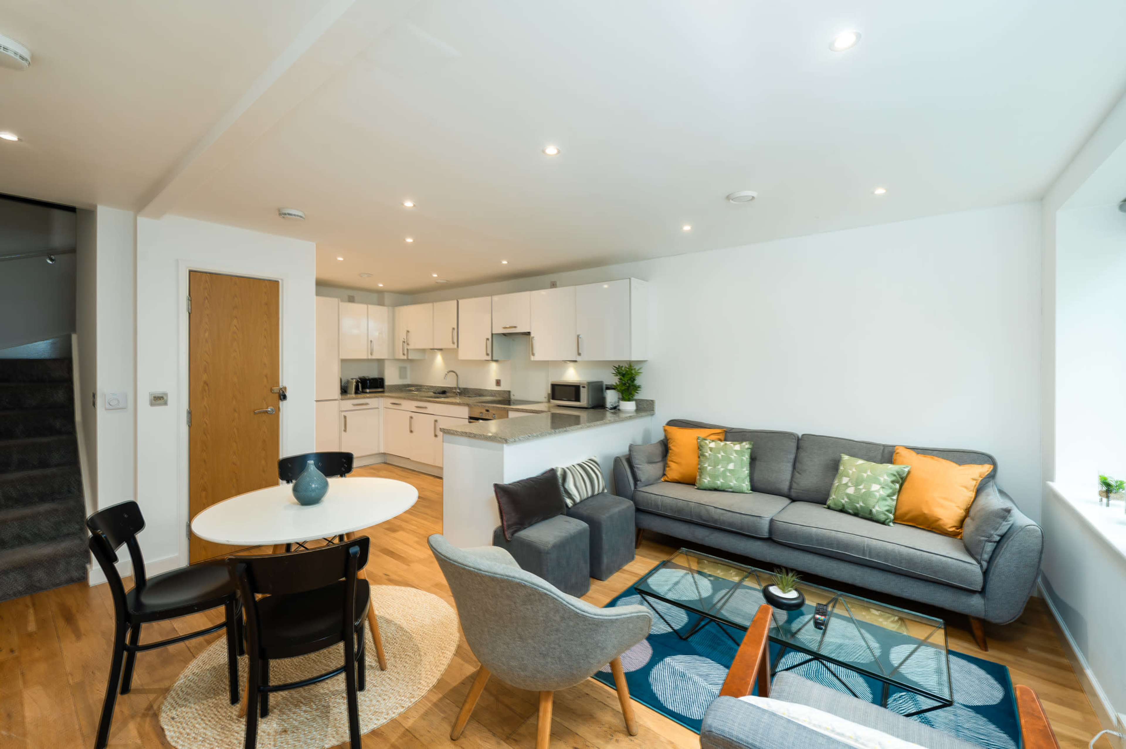 The image shows a modern living room with a gray sofa, a round dining table, and a kitchenette in a brightly lit open-plan space.