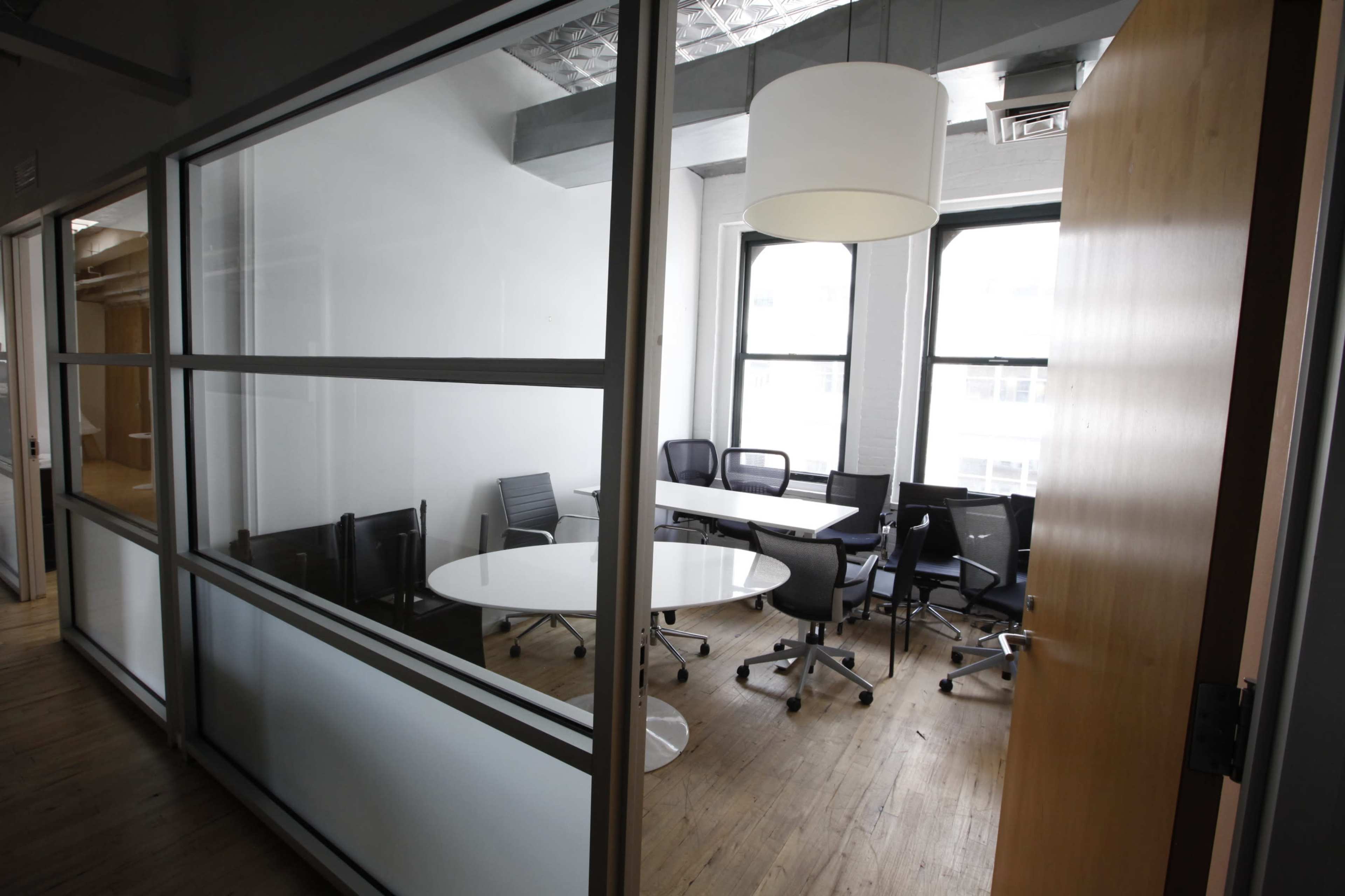 A modern office meeting room with a large round table surrounded by several chairs, visible through glass walls.