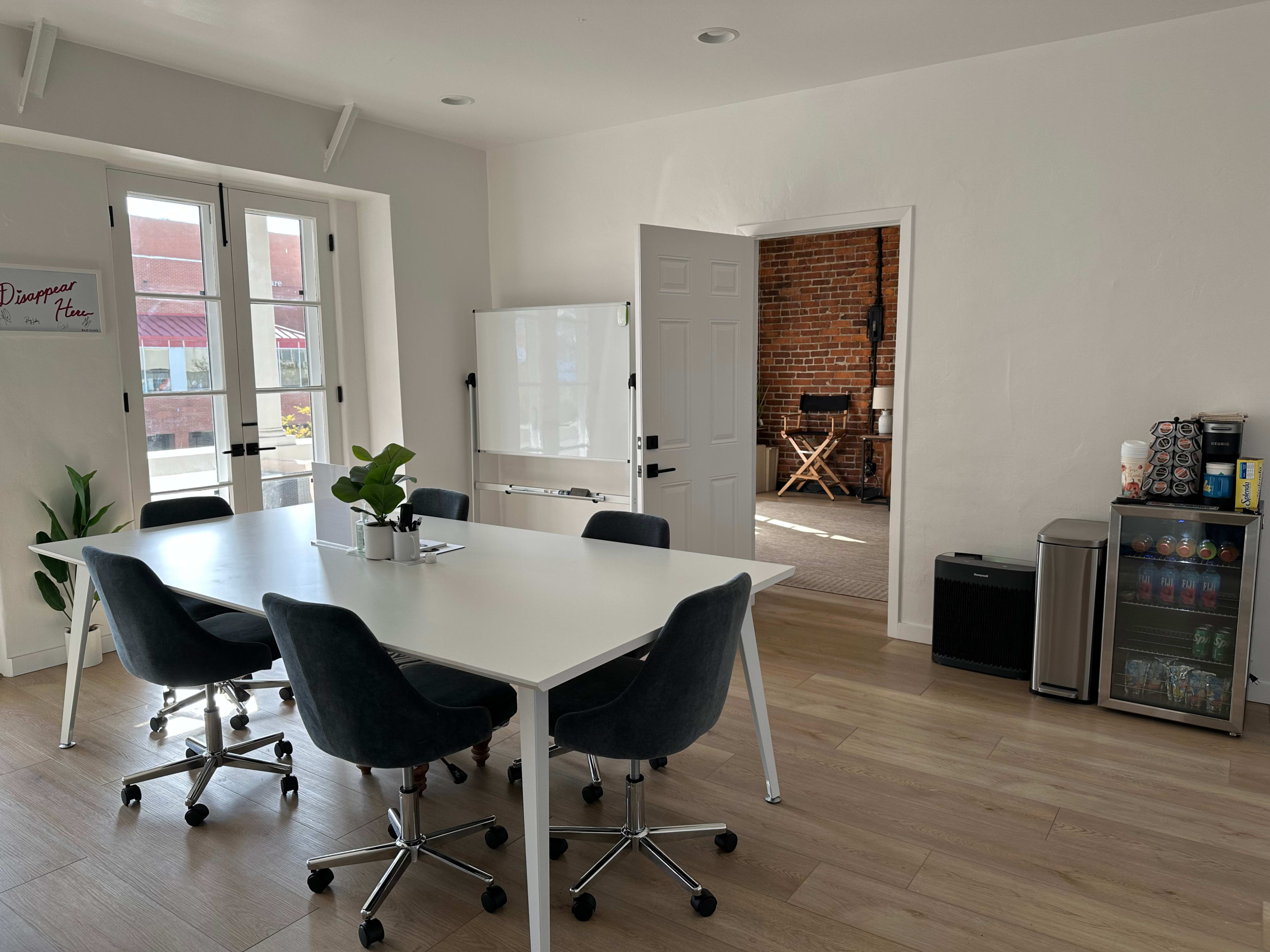 The image shows a modern office space with a large white table and gray chairs, a whiteboard, and a kitchenette displaying a small fridge and drinks.