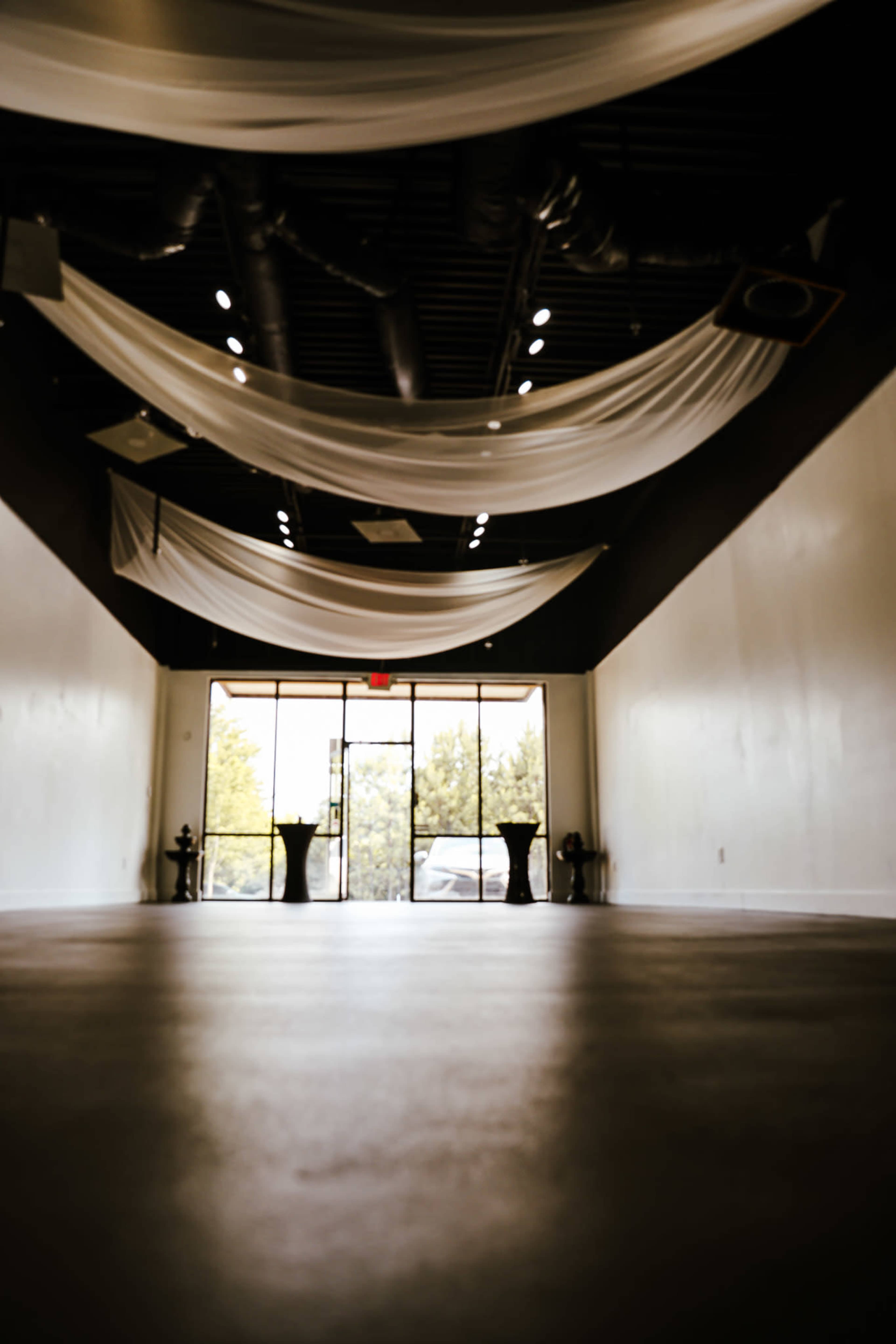 The image shows an empty indoor space with draped white fabric on the ceiling and large glass doors leading to the outside.