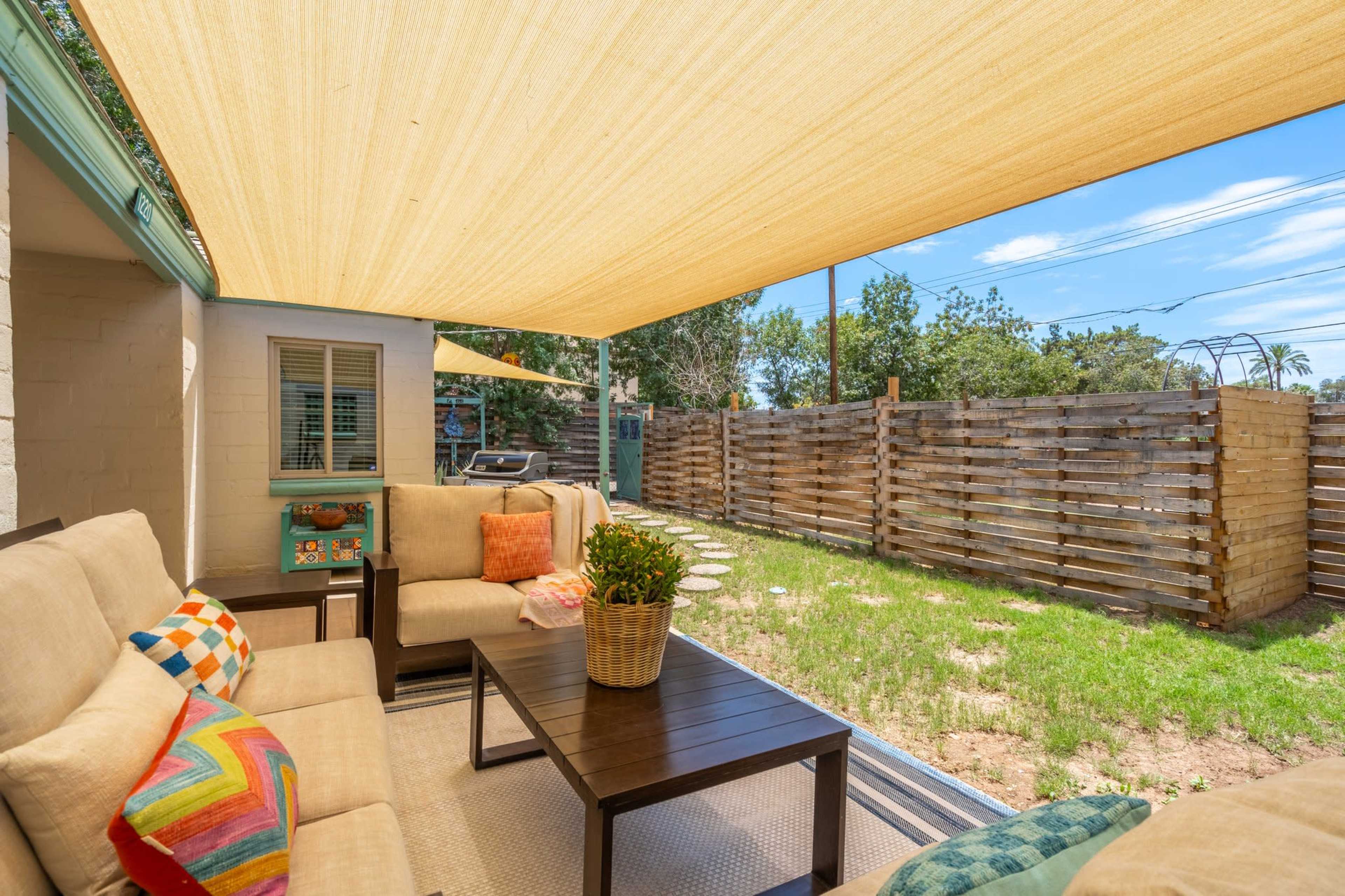 A patio area features a seating arrangement with cushions, a table, and a shaded awning in a backyard with grass and a wooden fence.