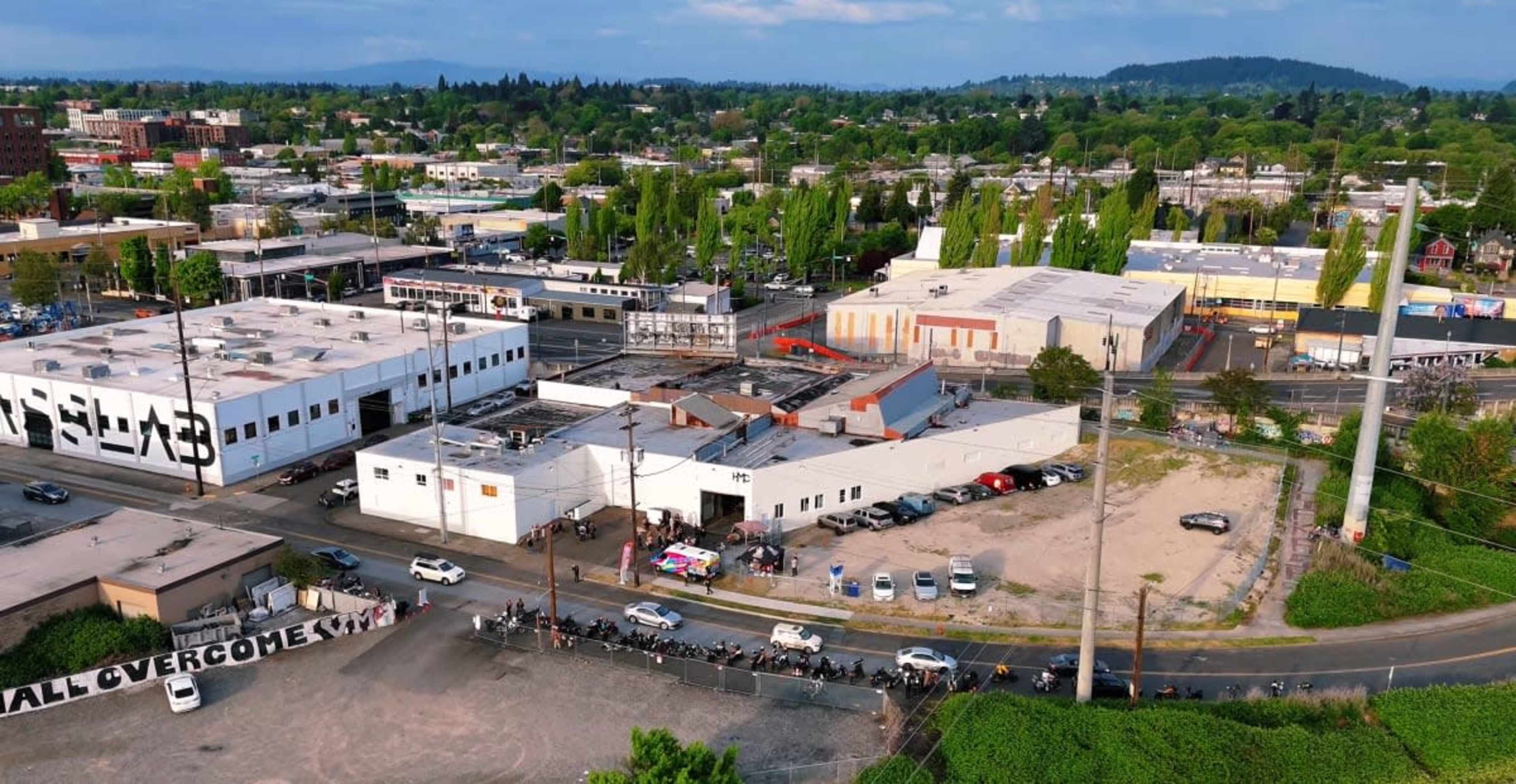 The image shows an aerial view of a cityscape with several buildings, parked cars, and a visible line of people outside one of the structures.