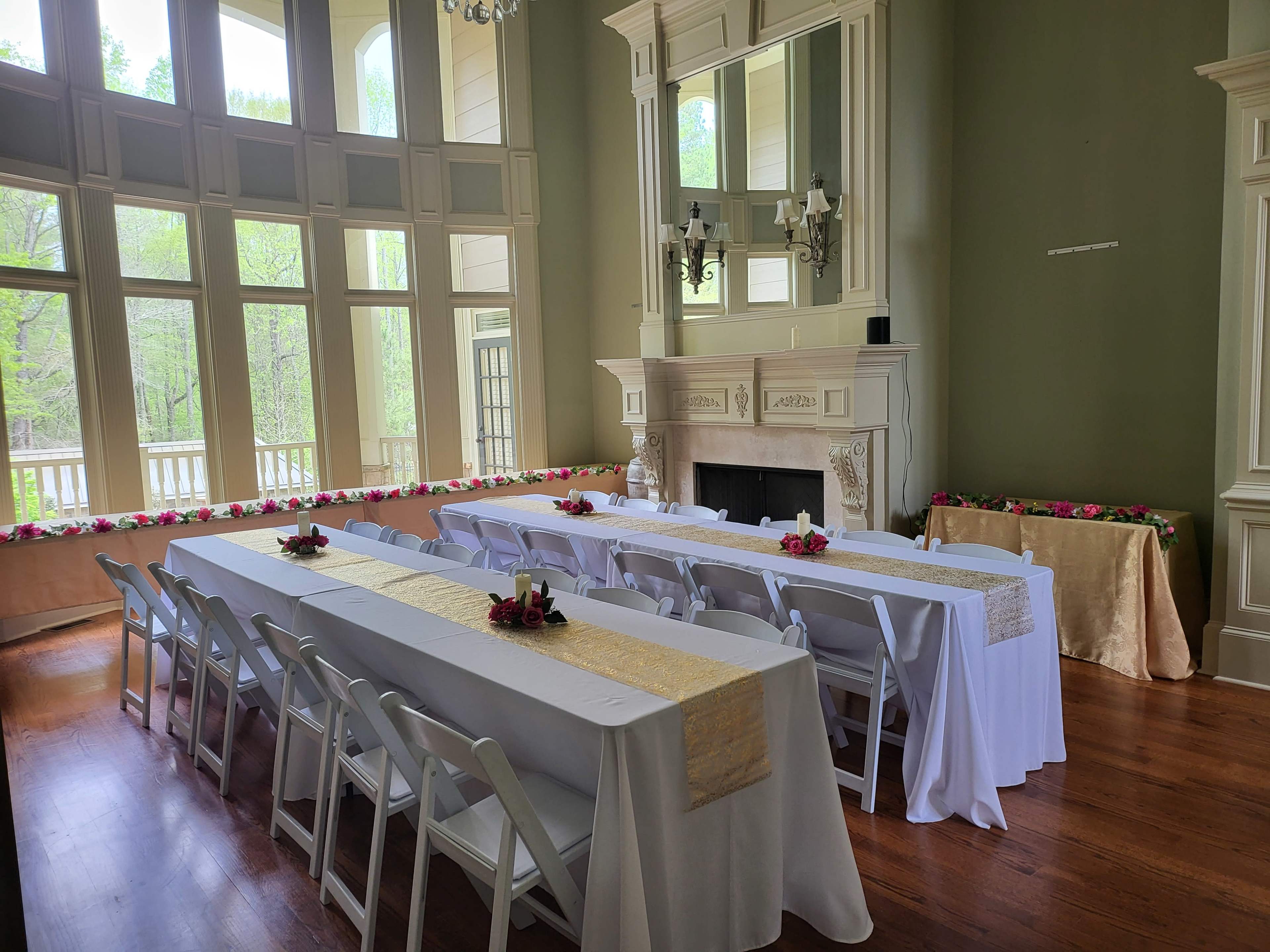 A large, elegantly arranged room features several tables covered with white linens, set up for an event, with floral decorations along the walls and near the fireplace.