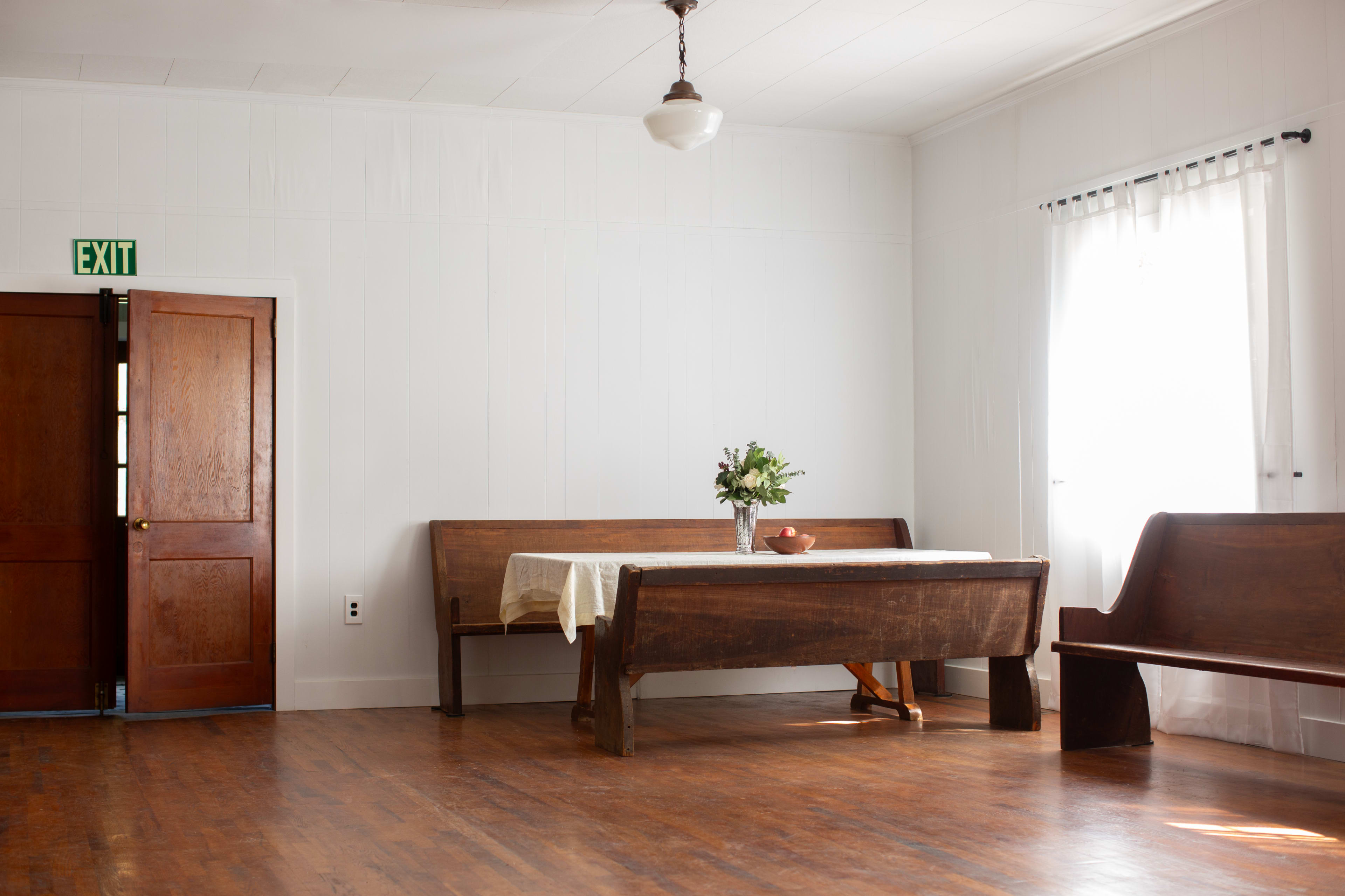 A simple room features a wooden table with a vase of flowers, benches along the walls, and a door leading outside.