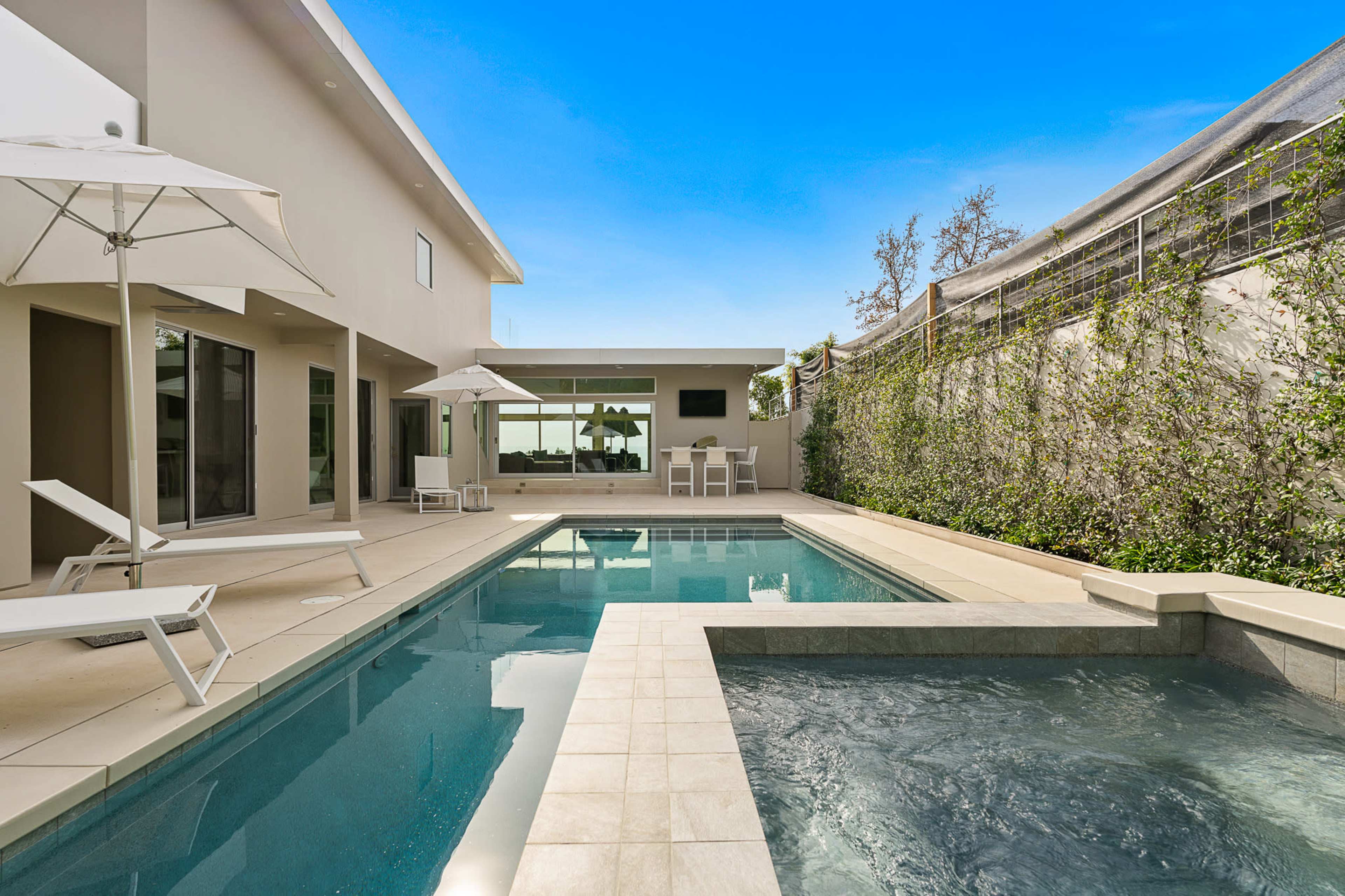 The image shows a modern outdoor pool area with a hot tub, lounge chairs, and umbrellas surrounded by a landscaped wall.