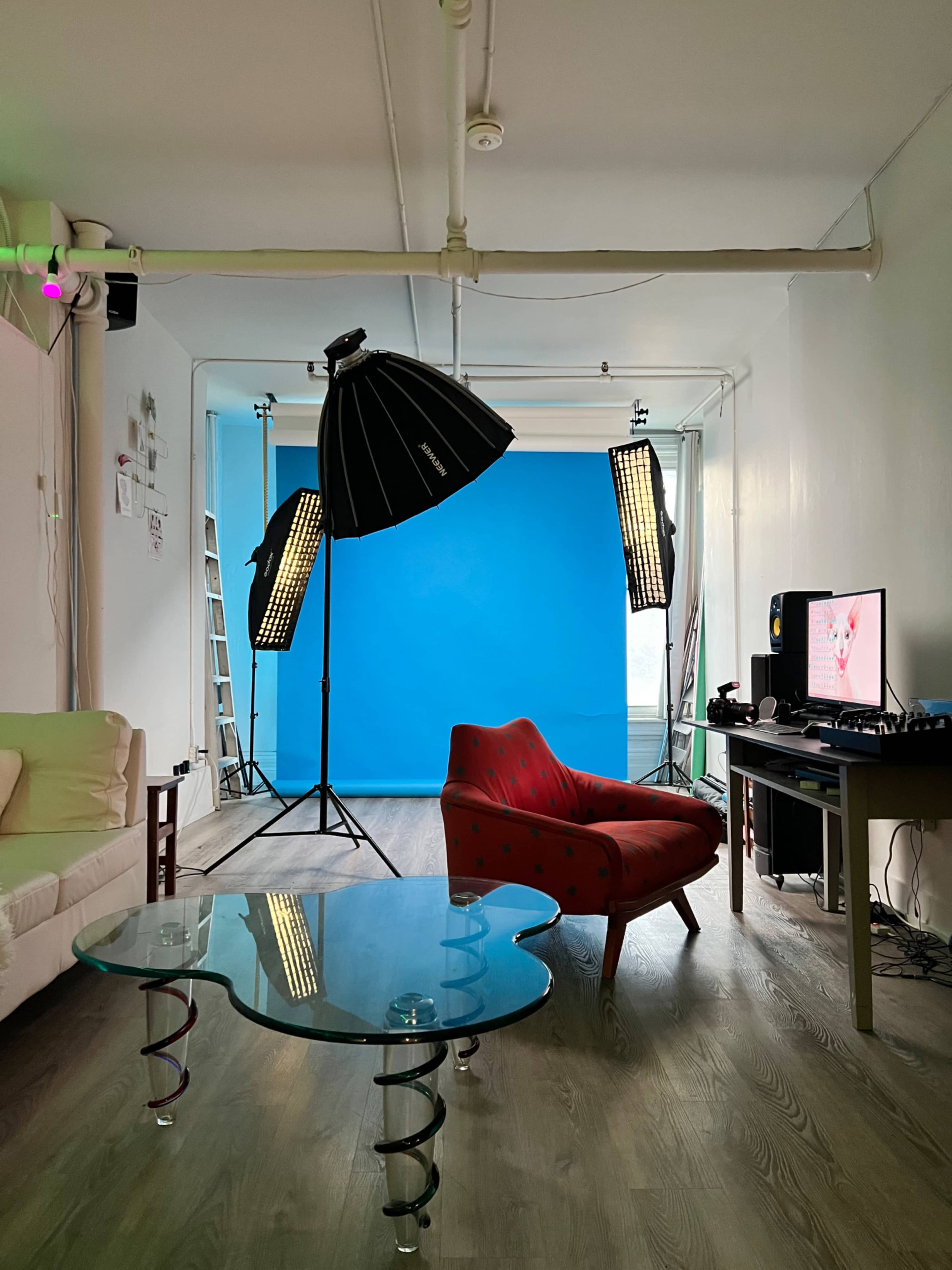 A photography studio features a red chair, a glass coffee table, and equipment including a blue backdrop and lighting stands.