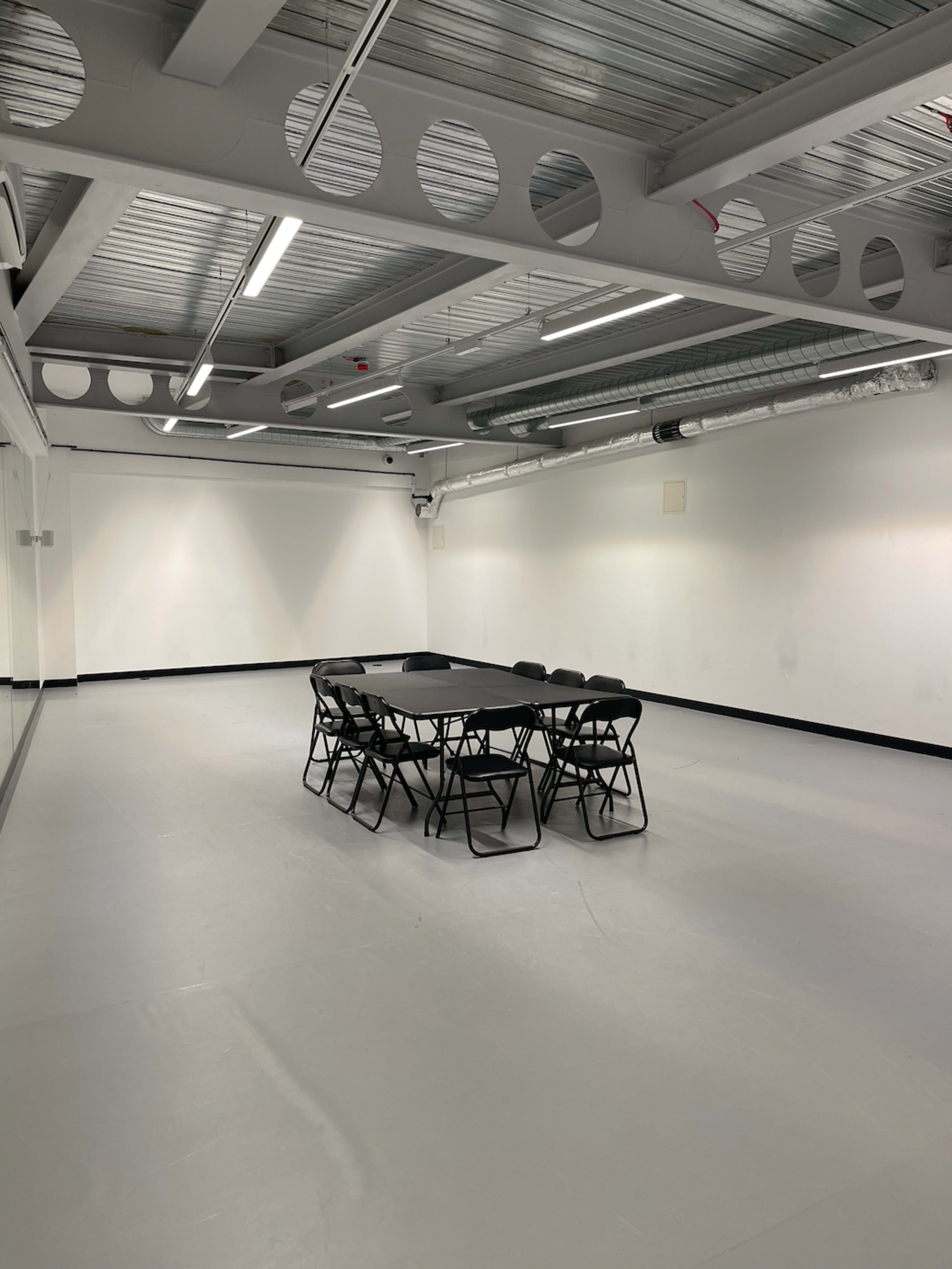 A large, empty room features a round table surrounded by black folding chairs on a smooth, gray floor.