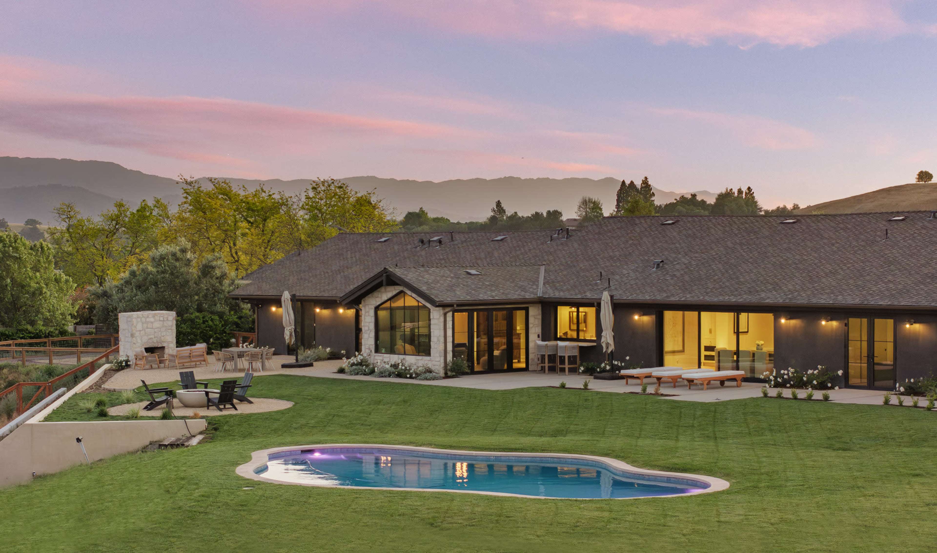 The image shows a modern single-story house with large windows, a swimming pool, and a landscaped yard, set against a backdrop of mountains and a colorful sky.