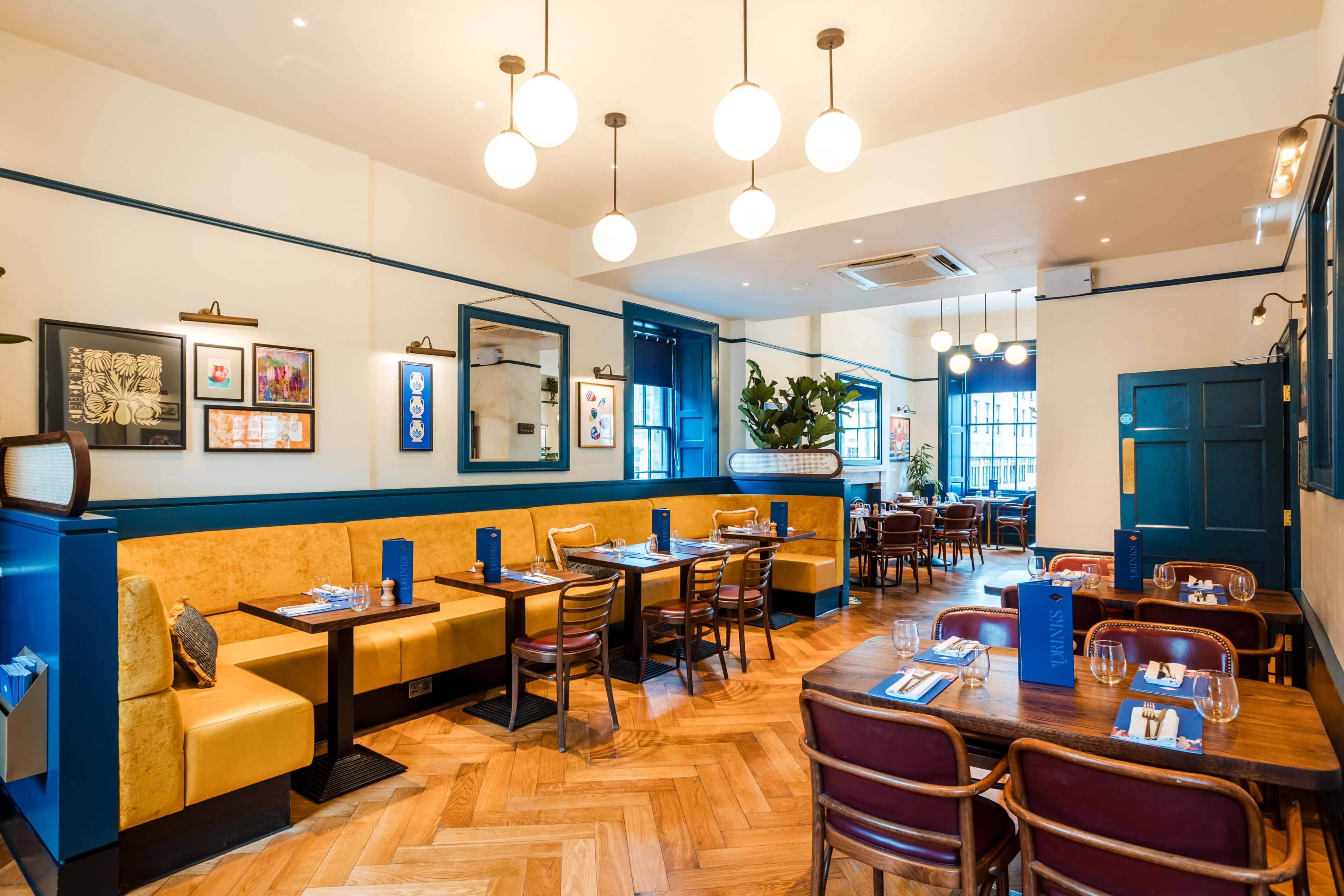 The image shows a well-lit restaurant interior featuring wooden floors, blue and yellow decor, and tables set with blue napkins and glassware.