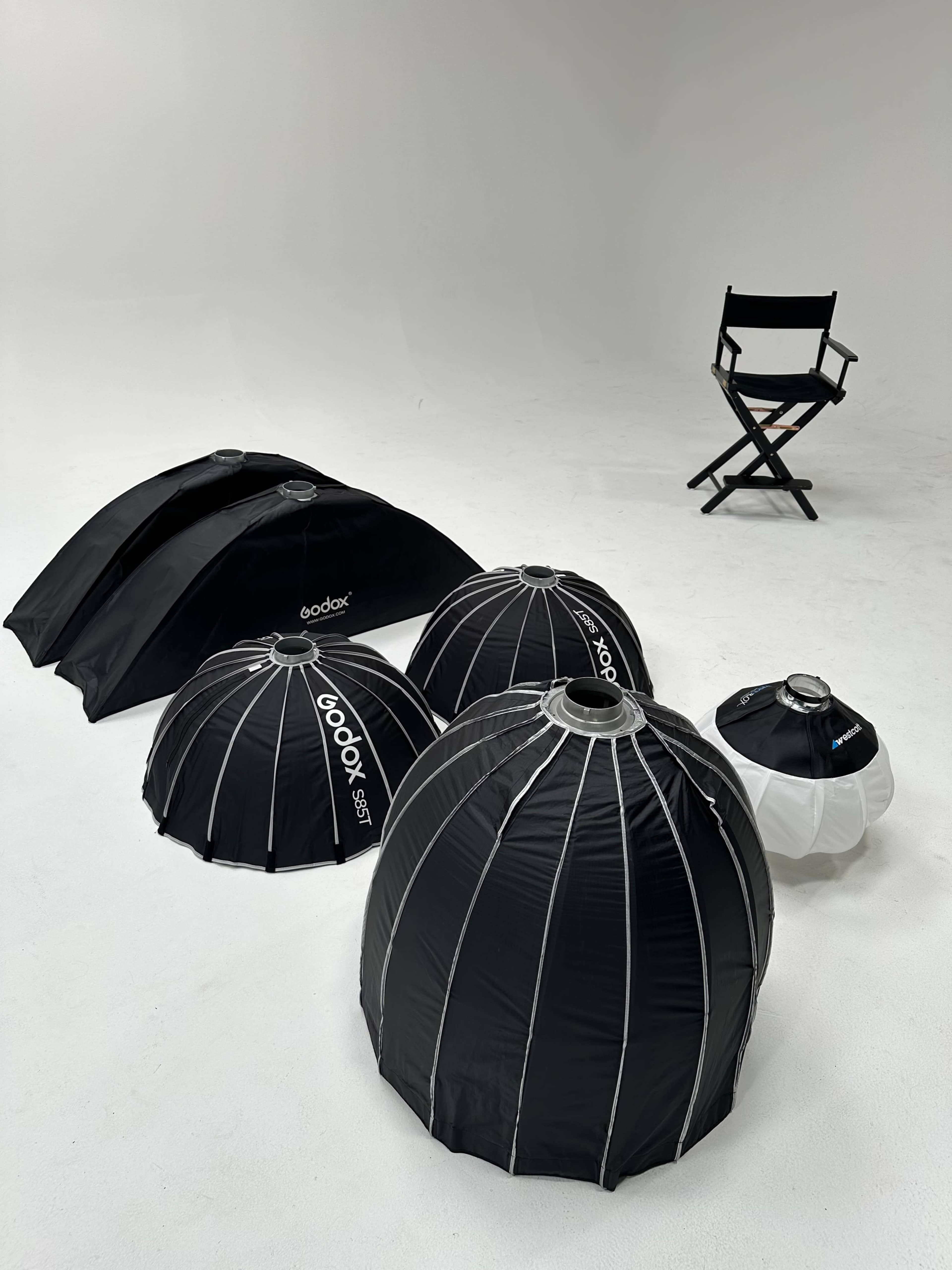 The image shows a collection of black and white softboxes arranged on a white studio floor, with a director's chair in the background.