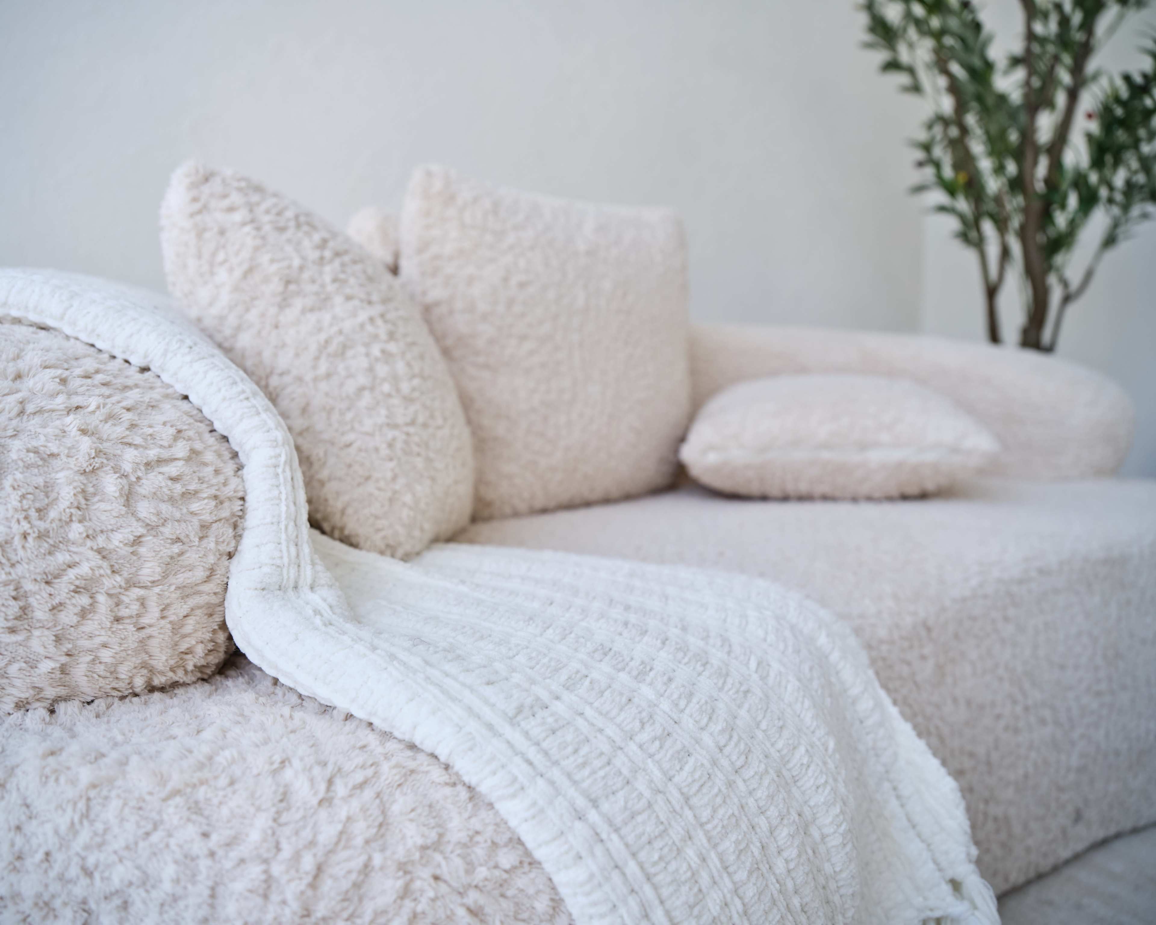 A cozy, textured light-colored sofa is adorned with multiple soft pillows and a white blanket draped over the arm.