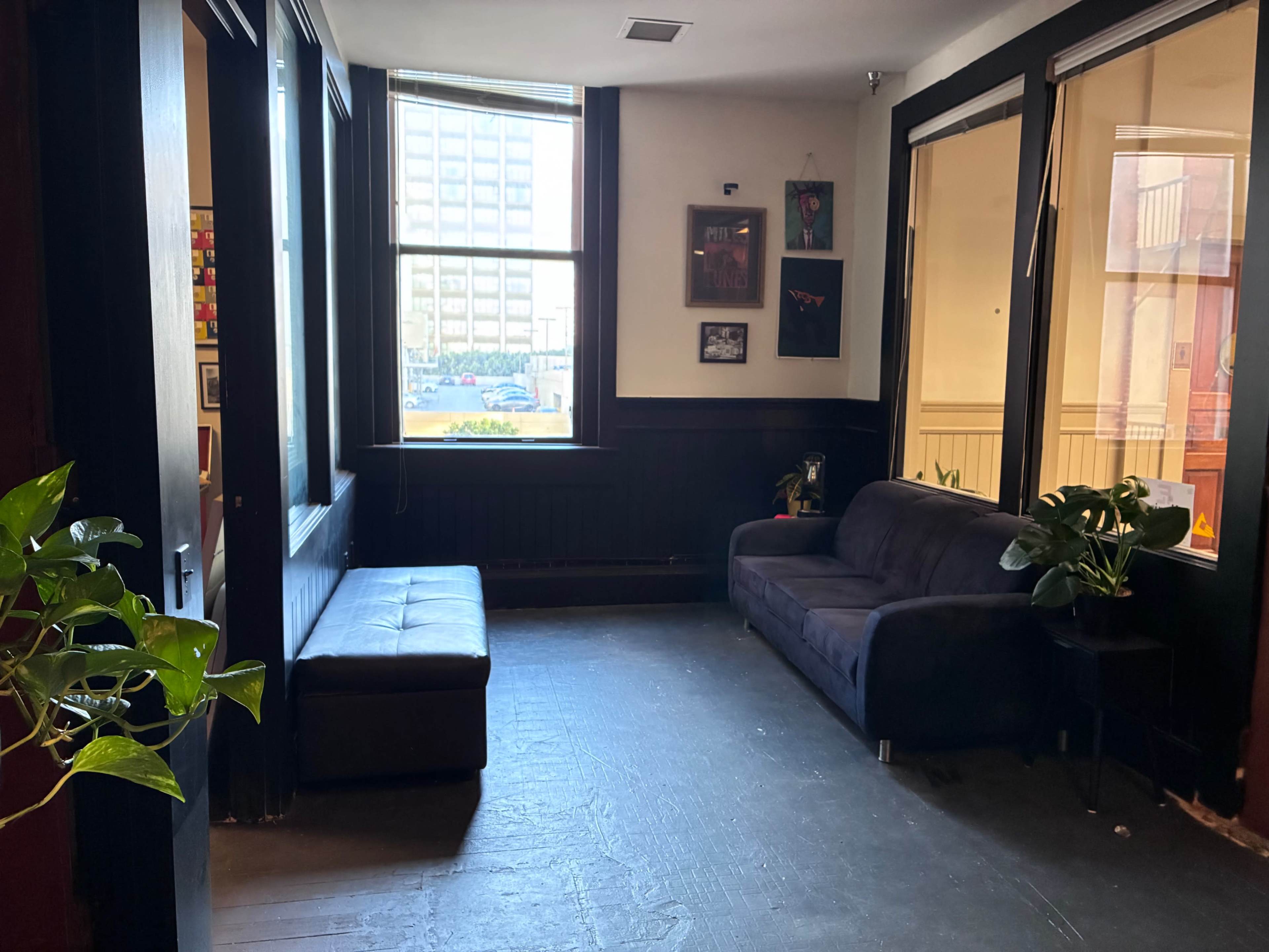The image shows a small interior seating area featuring a dark couch, a bench, and large windows allowing natural light to enter.
