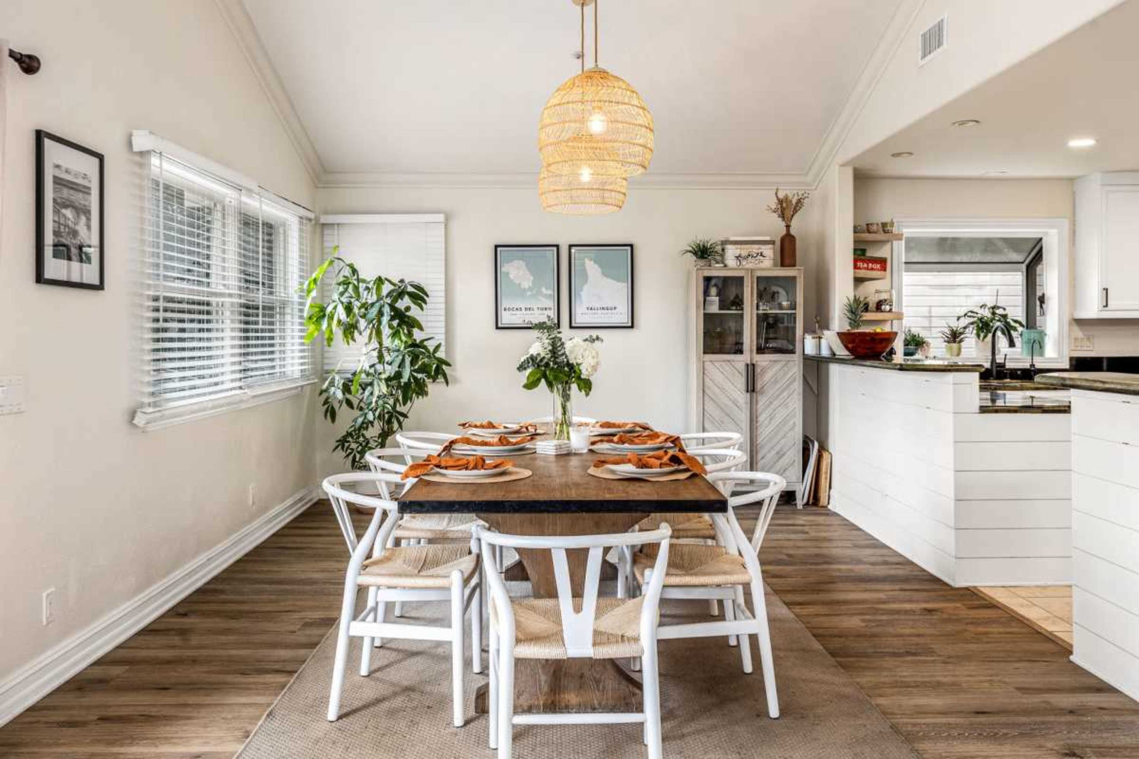 A dining area features a wooden table set for six, surrounded by white chairs, with a pendant light overhead and plants in the corners.