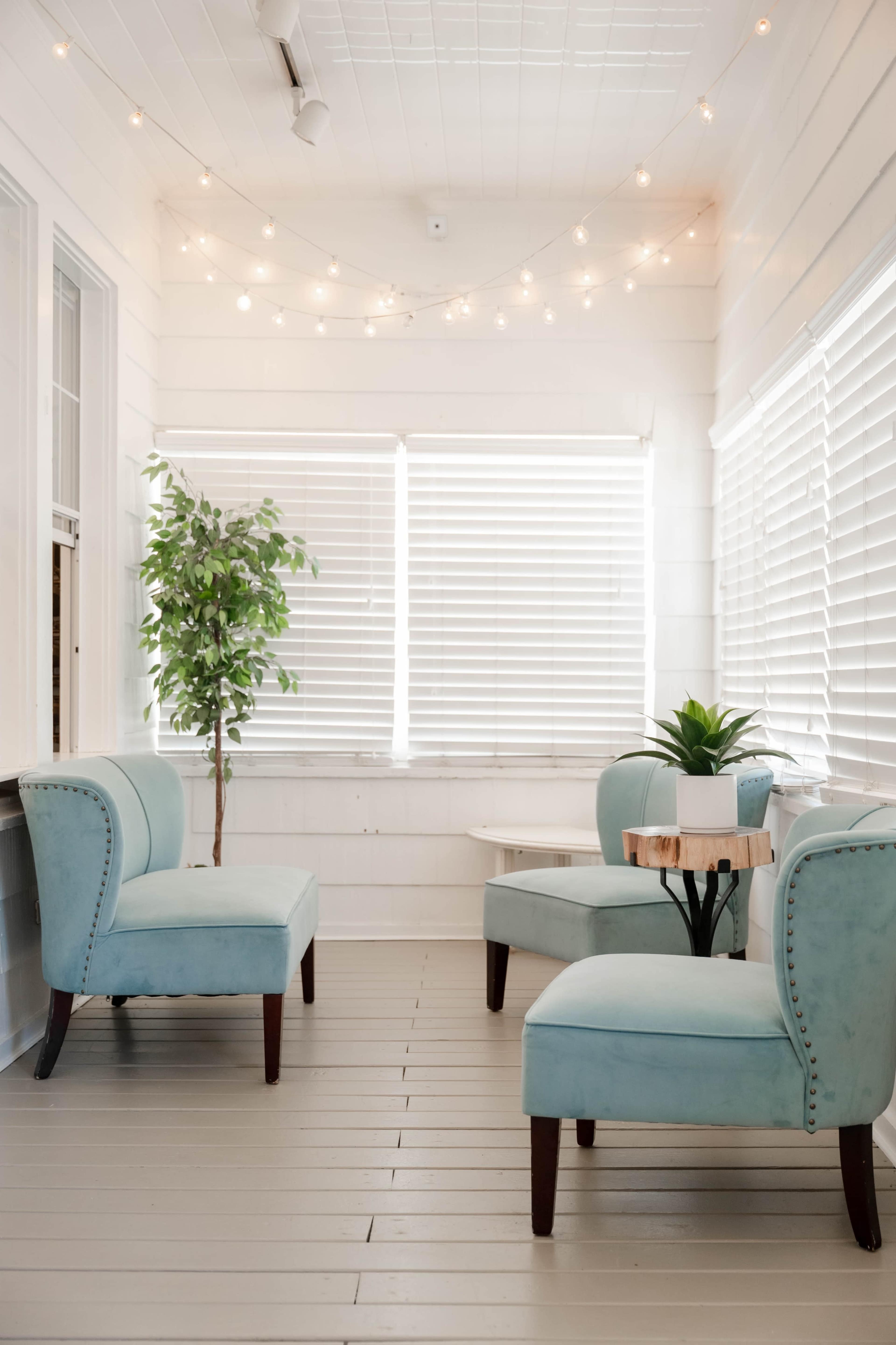 A bright, airy room features two turquoise chairs and a small table beside a potted plant, with string lights above and shutters covering the windows.