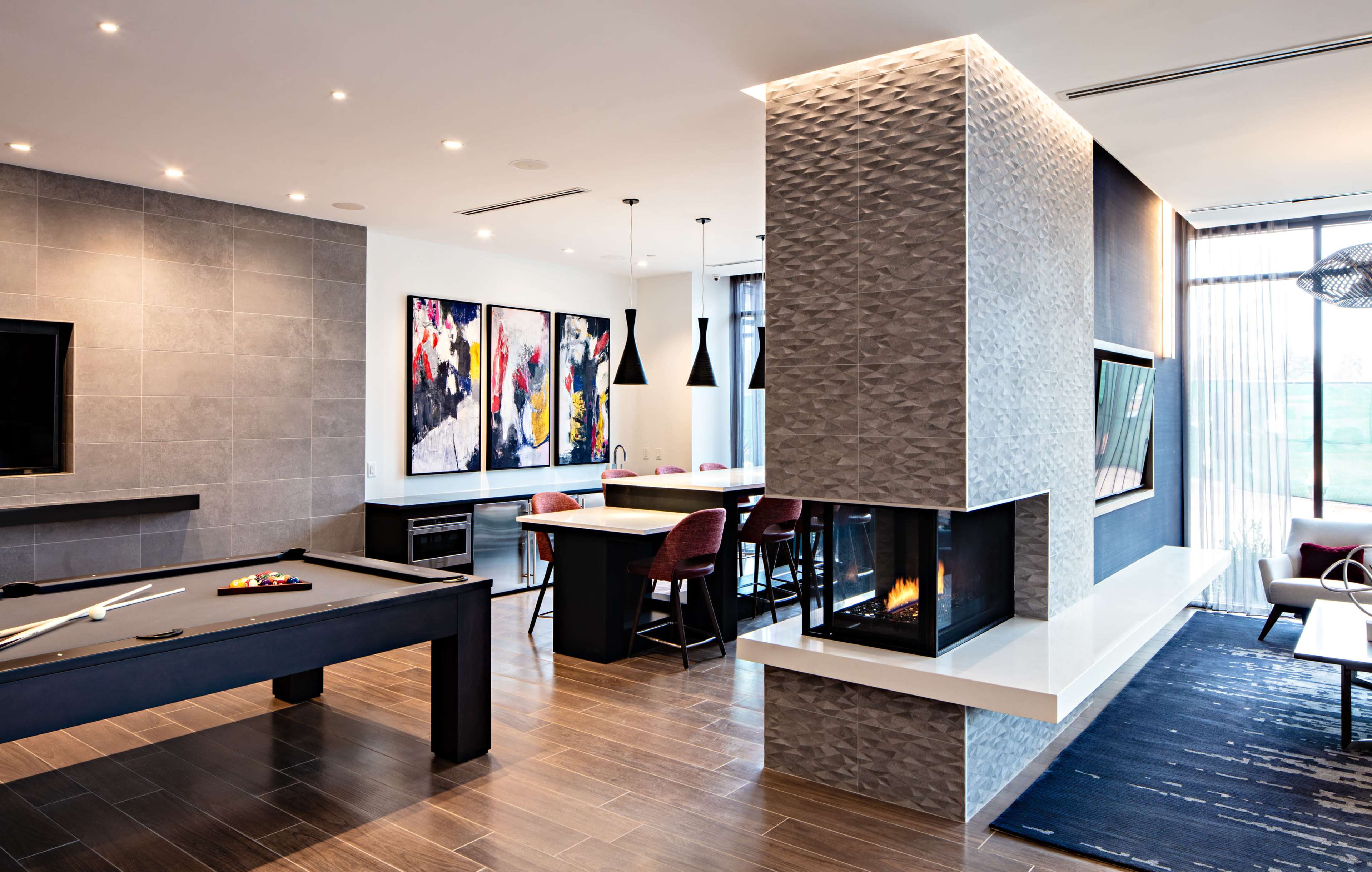A modern living space featuring a pool table, a dining area with bar stools, a two-sided fireplace, and large windows with natural light.