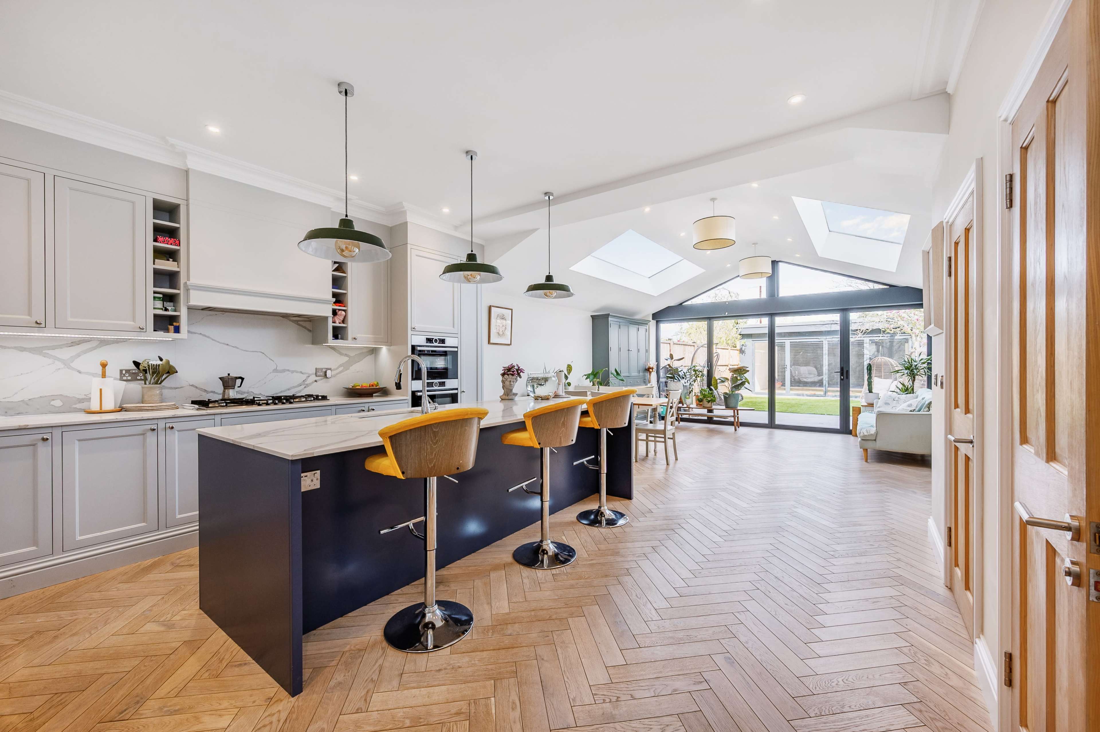 A modern kitchen features a central island with bar stools, sleek cabinetry, and large windows overlooking a backyard patio.