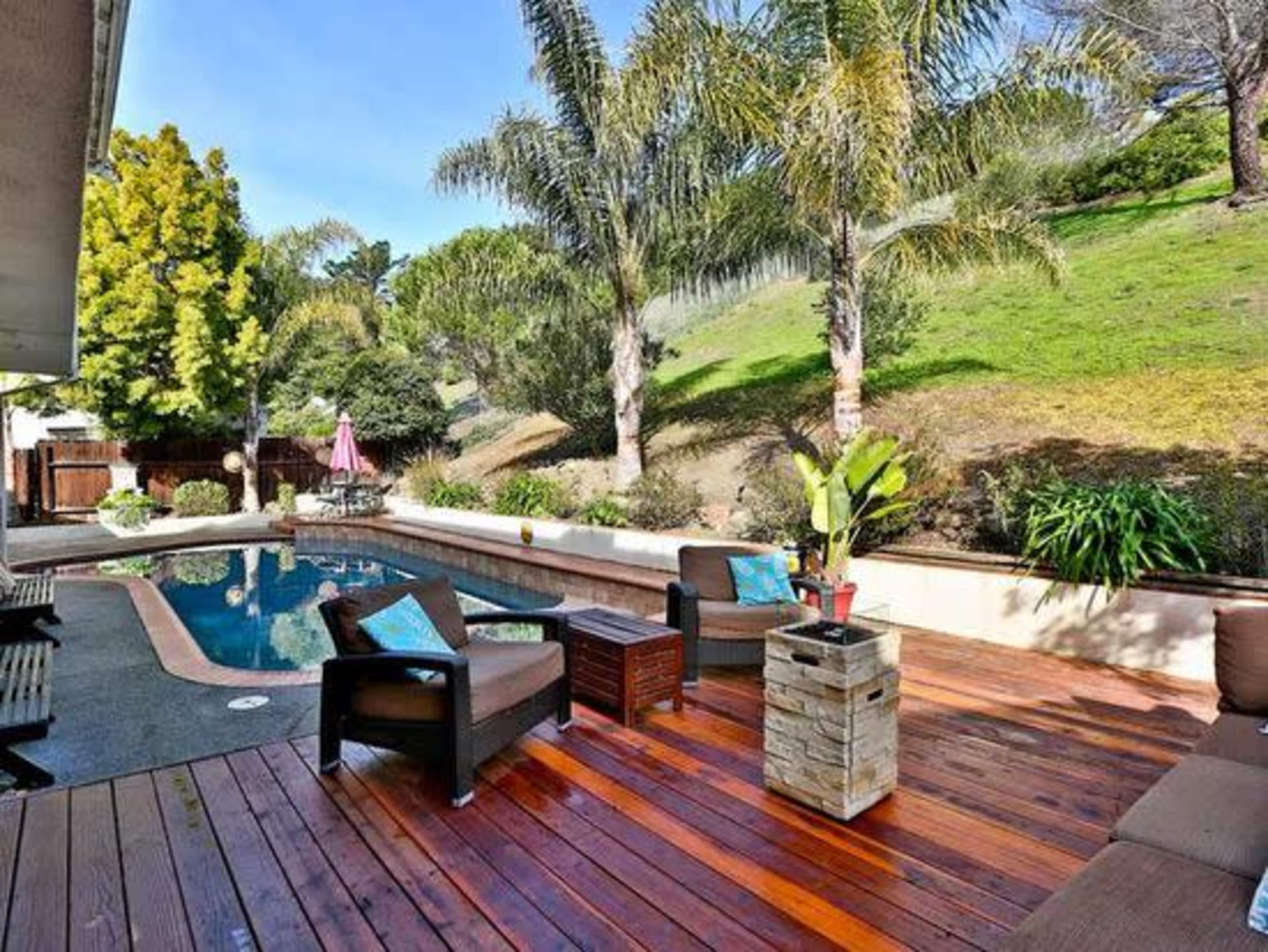 A backyard patio features a swimming pool surrounded by palm trees and wooden furniture.