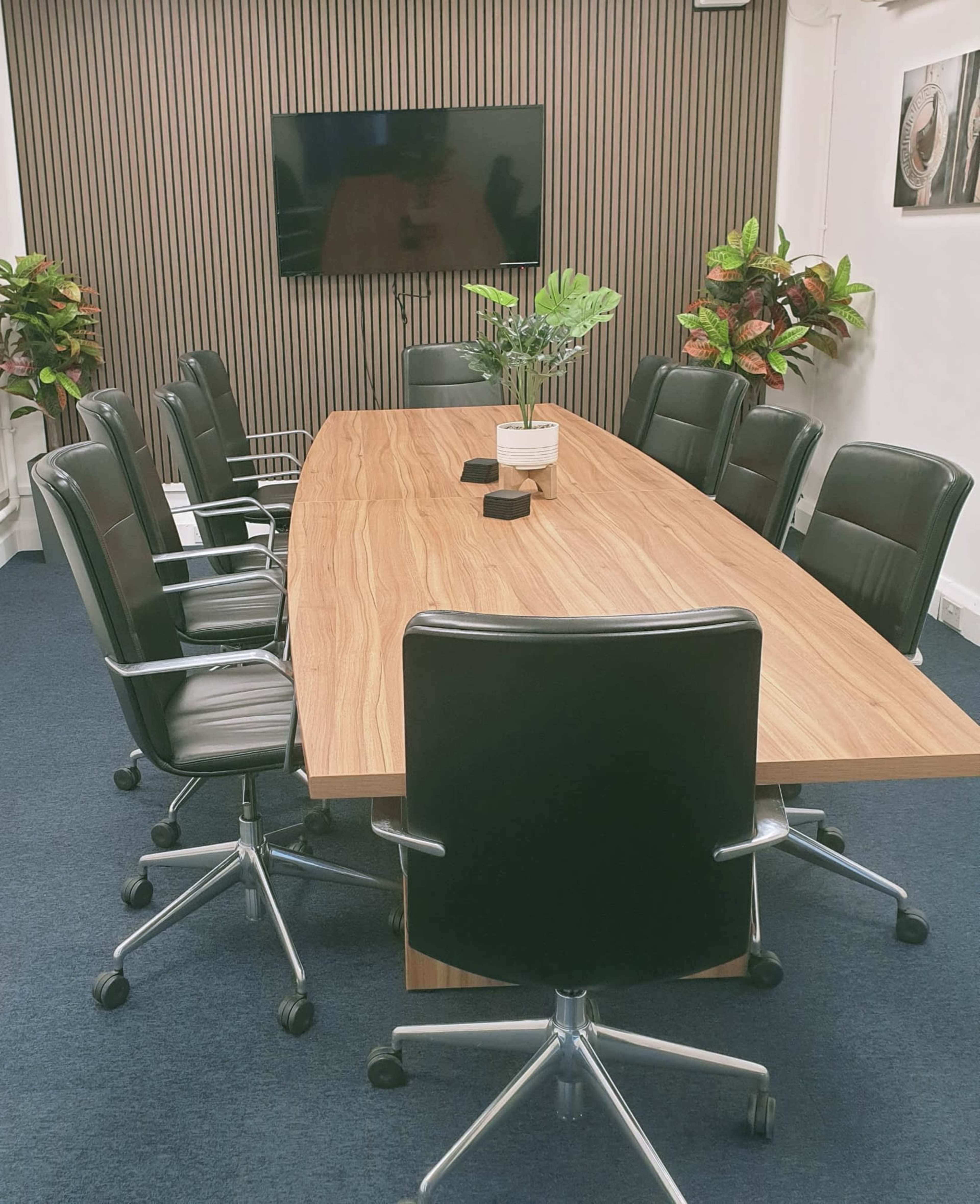 A large conference table with black ergonomic chairs surrounds it, and a television is mounted on a striped wall in a well-lit meeting room.