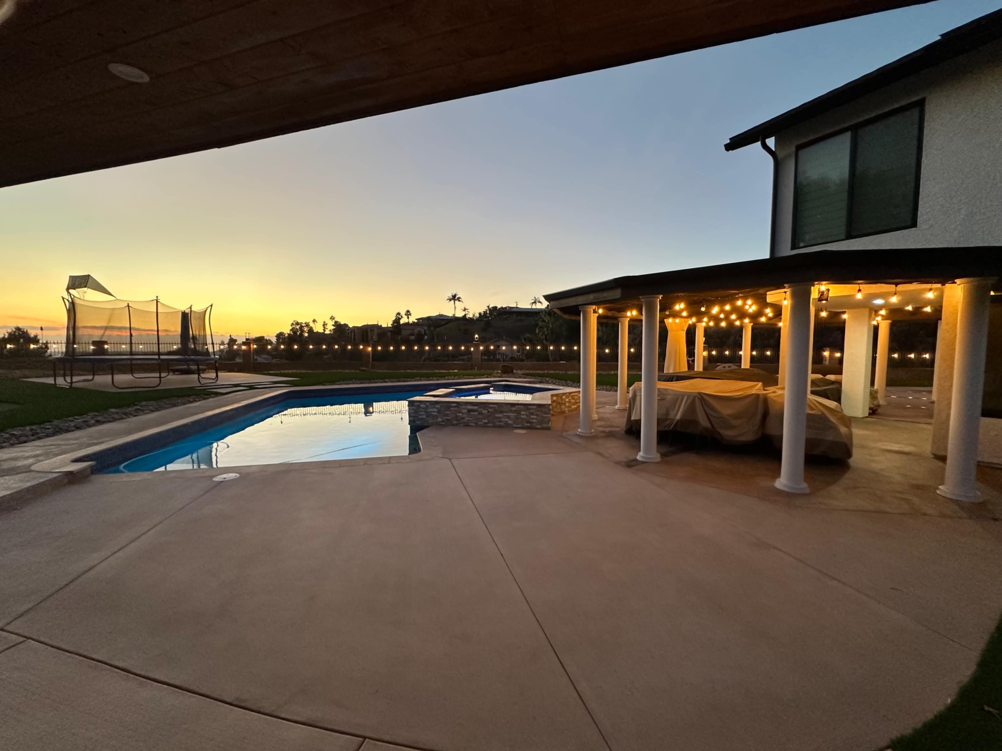 A backyard with a swimming pool, a spa, and a covered patio area, set against a backdrop of a sunset.