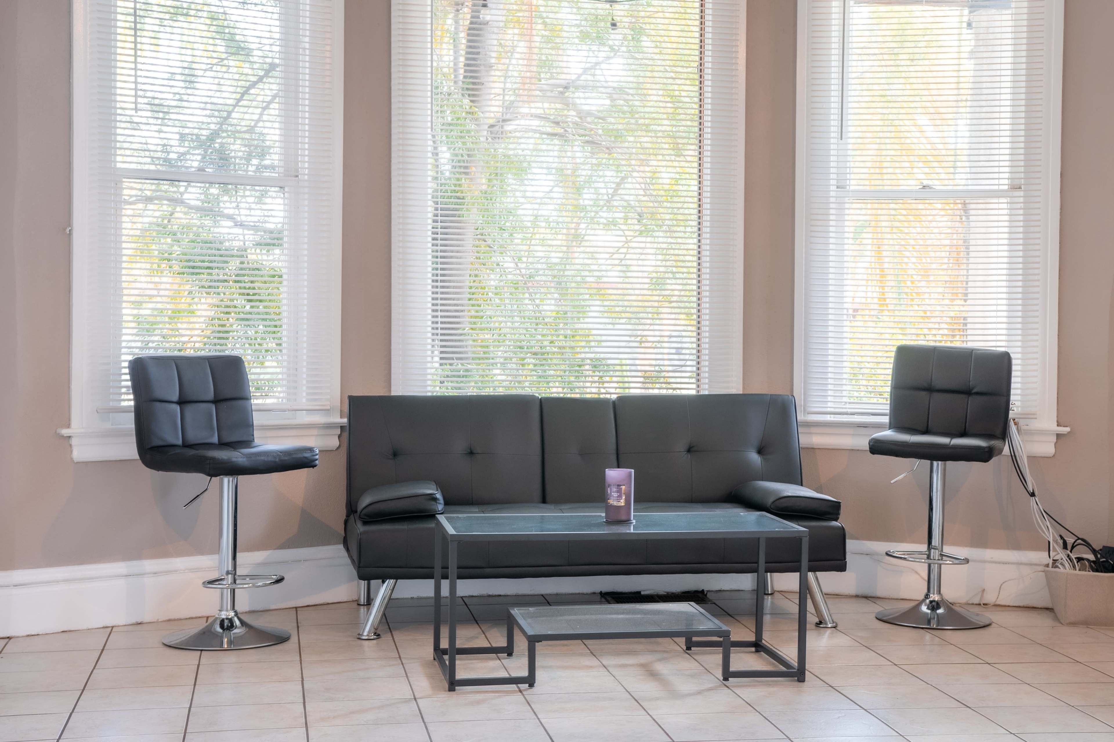 A modern living room features a black sofa flanked by two barstools, with a glass coffee table in front, all positioned near large windows.