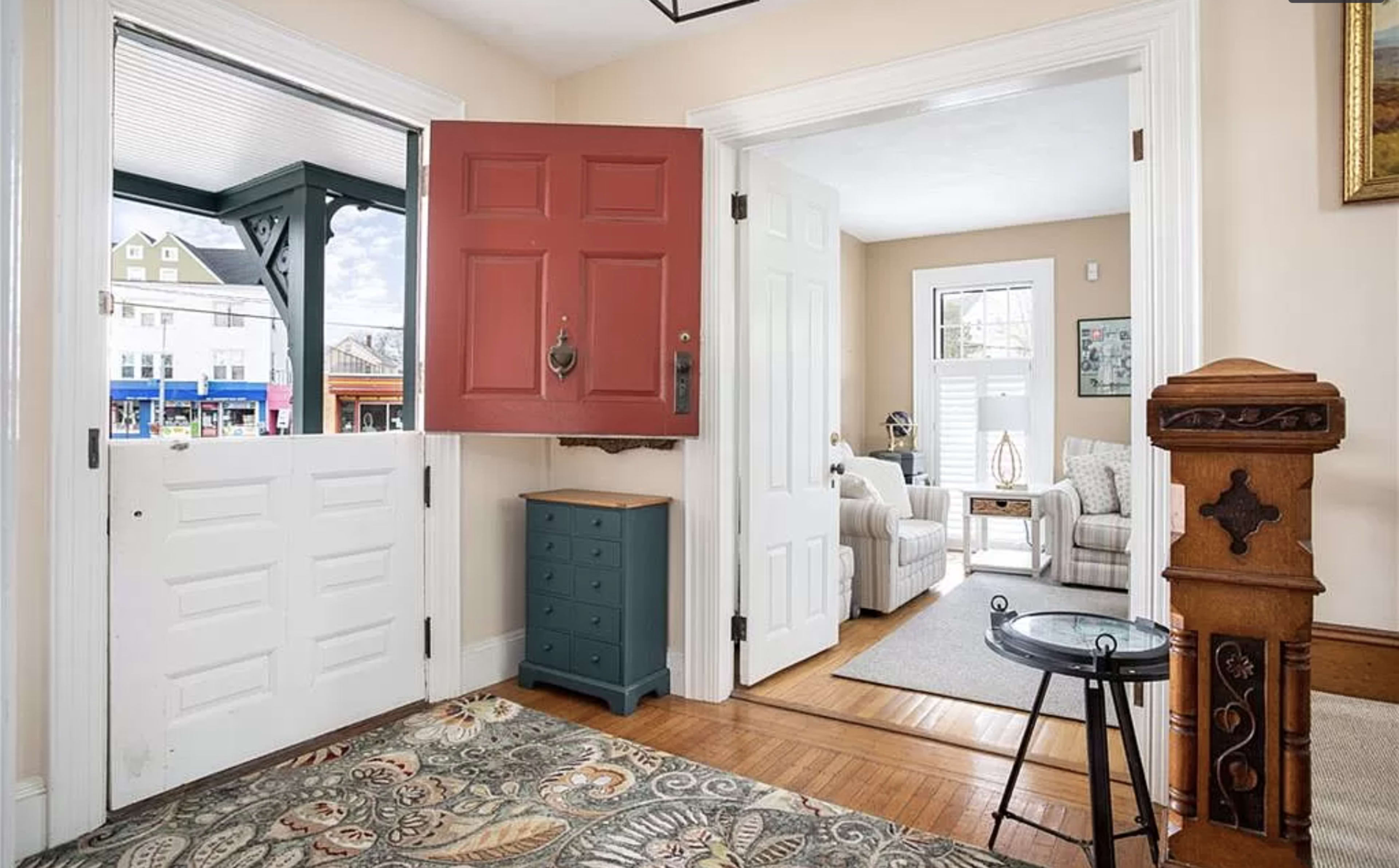 The image shows a bright entryway with a red front door open to a living room featuring two white armchairs and a side table, along with a view of a street outside.