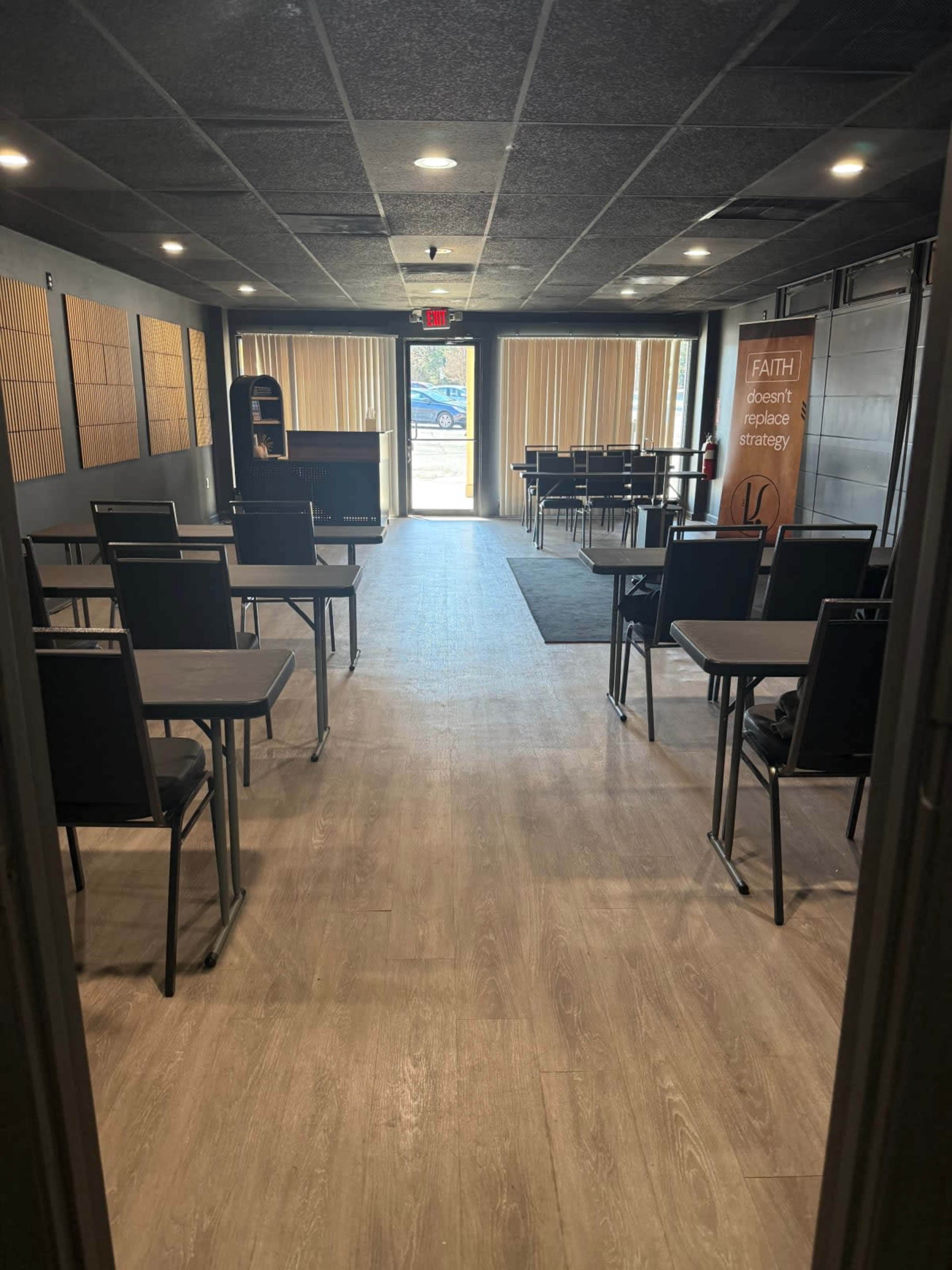 The image shows a spacious indoor room set up for a meeting, featuring several tables and chairs arranged in rows, with large windows allowing natural light to enter.