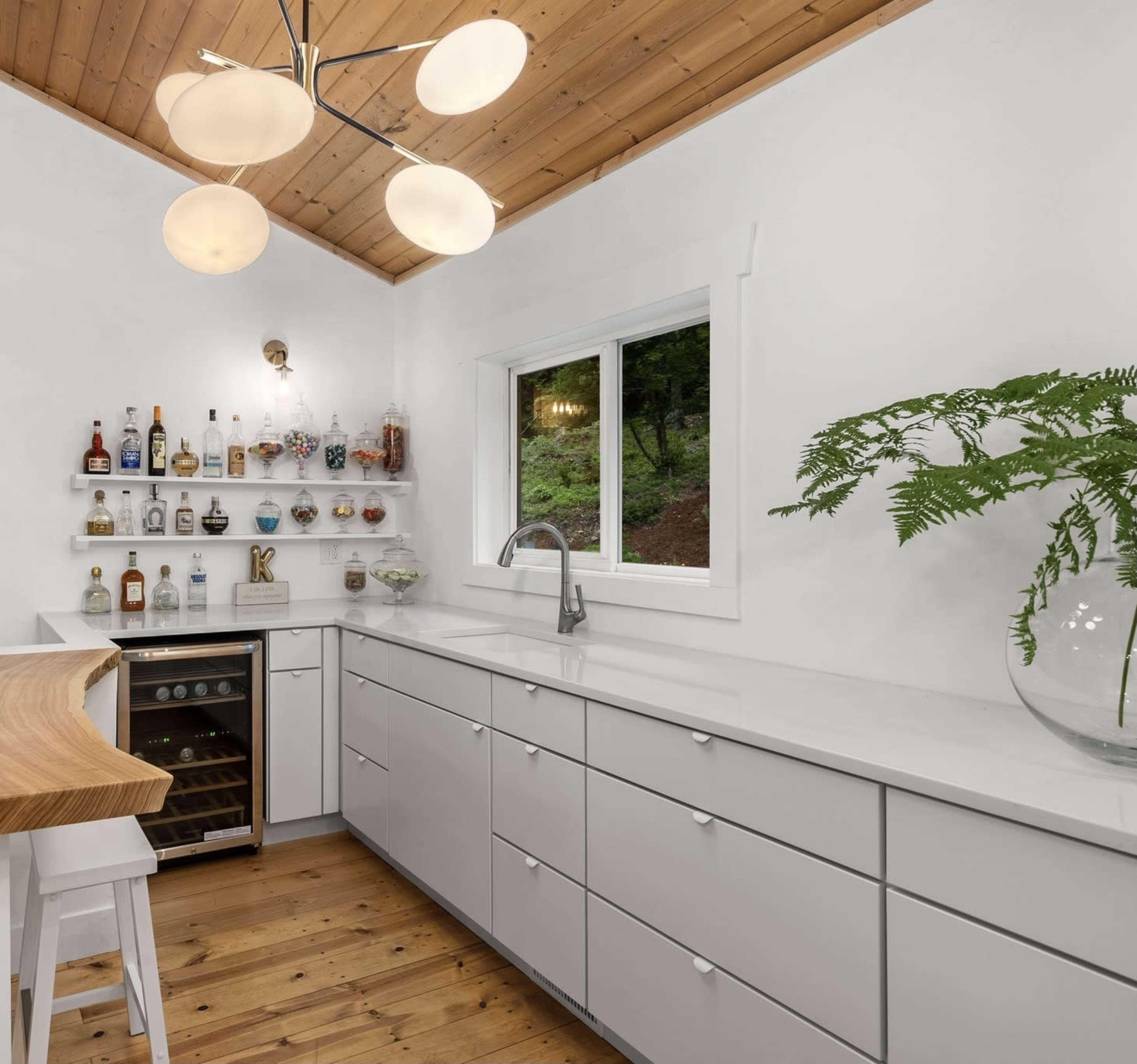 The kitchen features a modern design with a white countertop, wooden accents, and a variety of bottles displayed on a shelf.