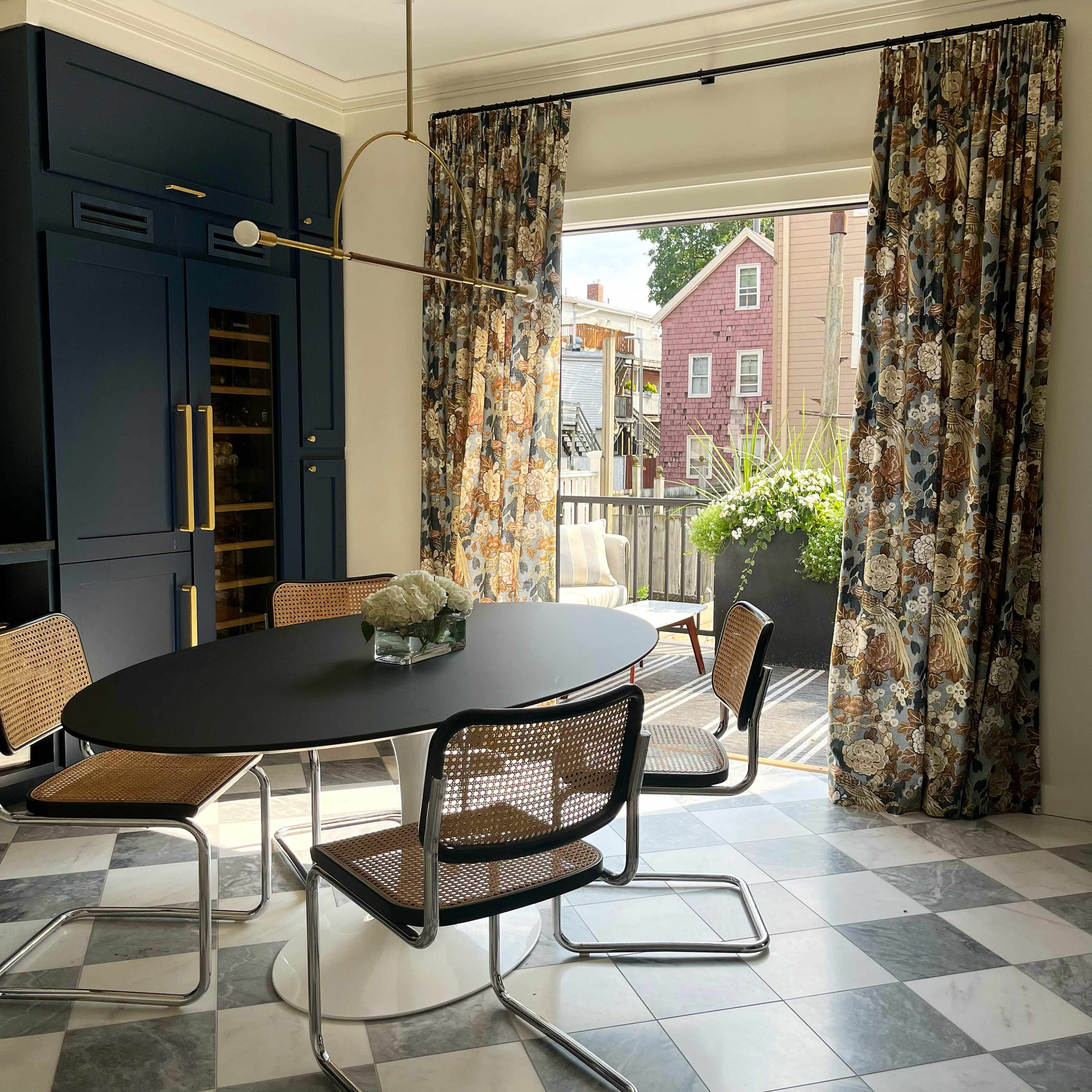 A modern dining area features a round table with four chairs, large patterned curtains, and a view of an outdoor space through a sliding door.
