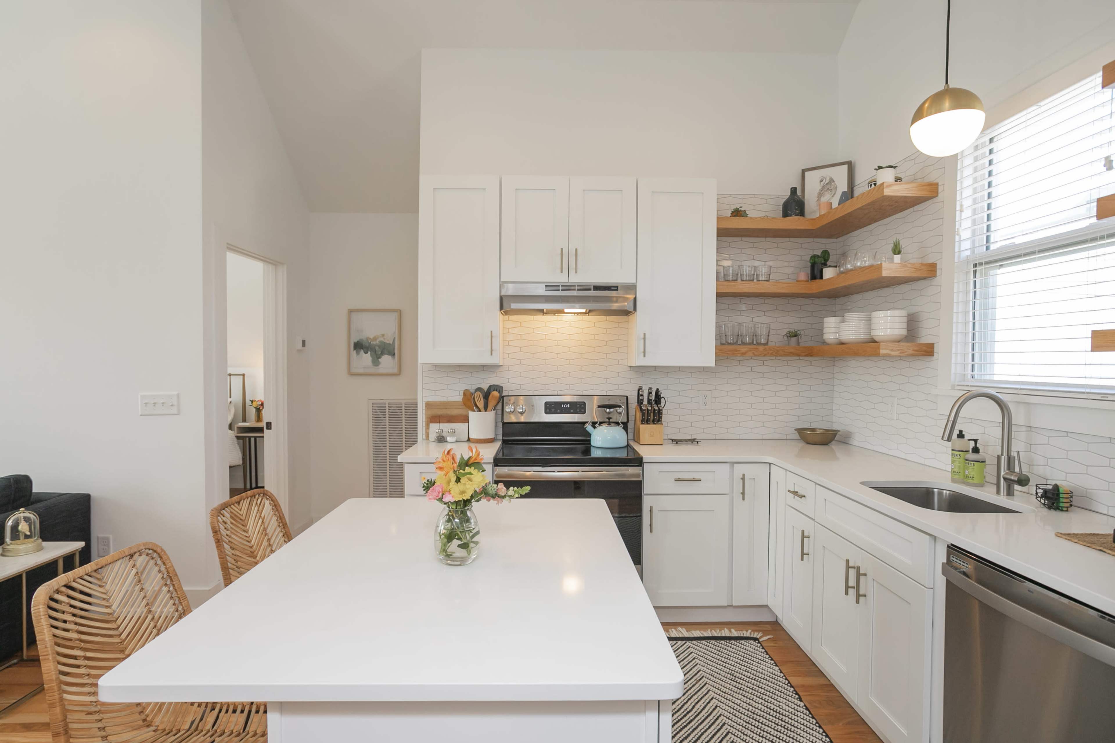 The image shows a modern kitchen with white cabinetry, open shelves displaying dishware, a central island with a vase of flowers, and stainless steel appliances.