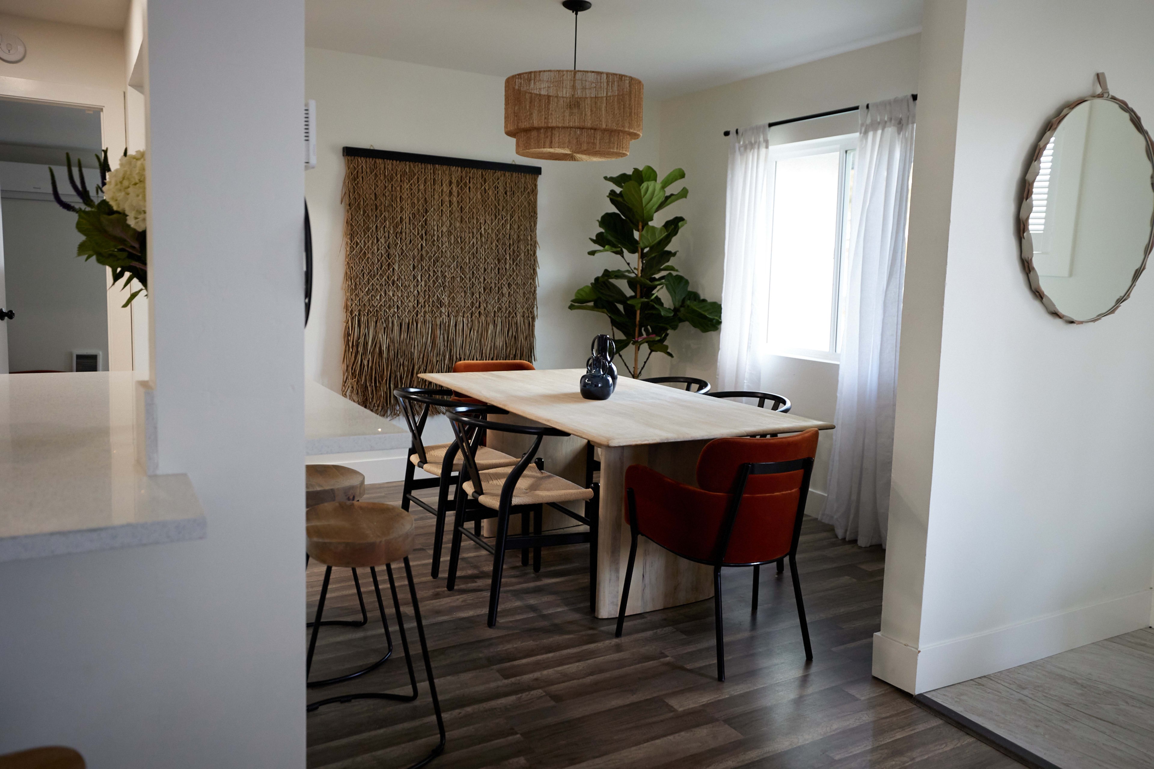 This image shows a modern dining area featuring a wooden table surrounded by black and red dining chairs, with a large wall hanging and greenery in the background.