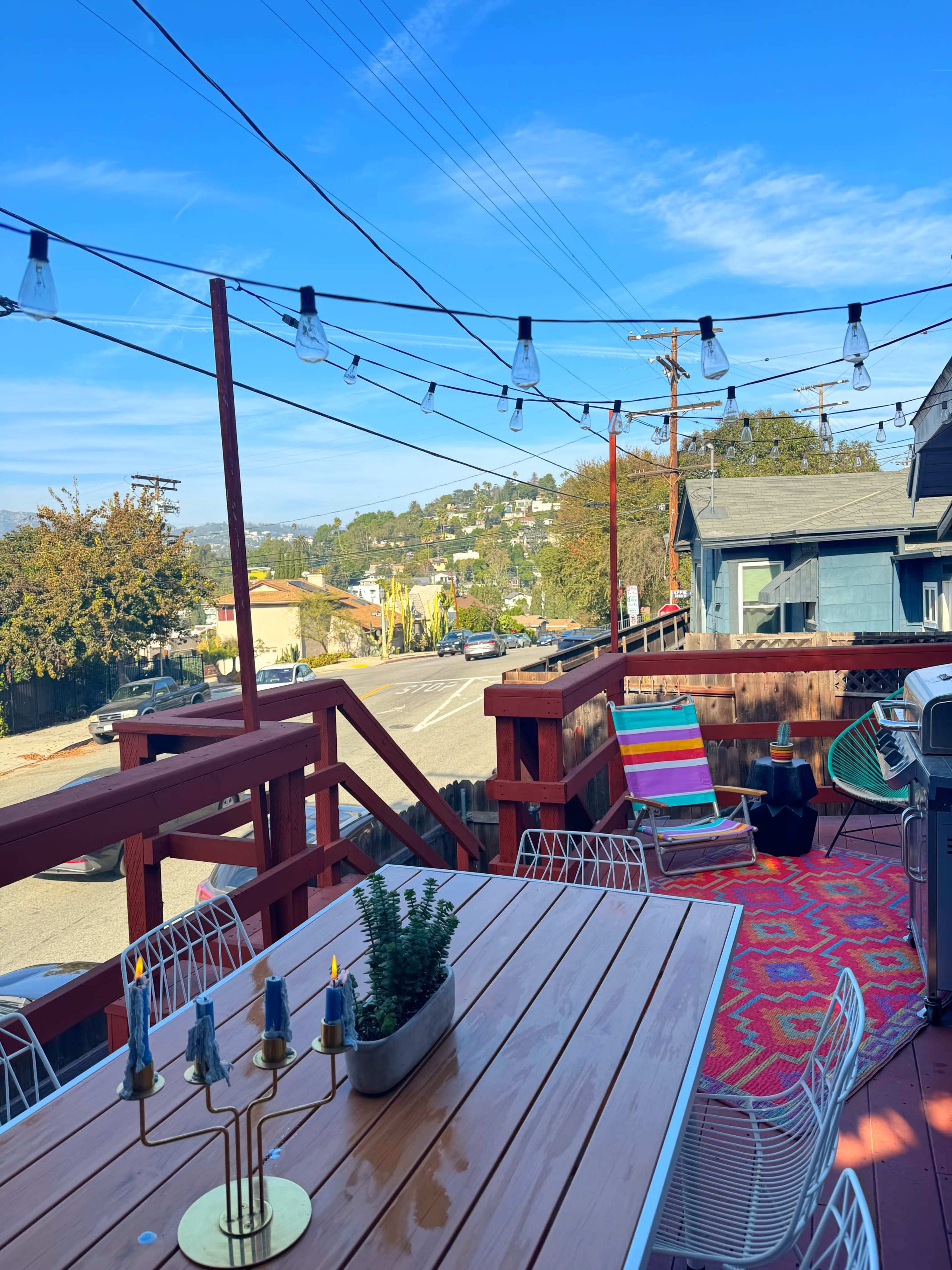 The image shows a wooden deck with patio furniture, a table with candles, and strings of lights, overlooking a neighborhood with houses and a clear blue sky.