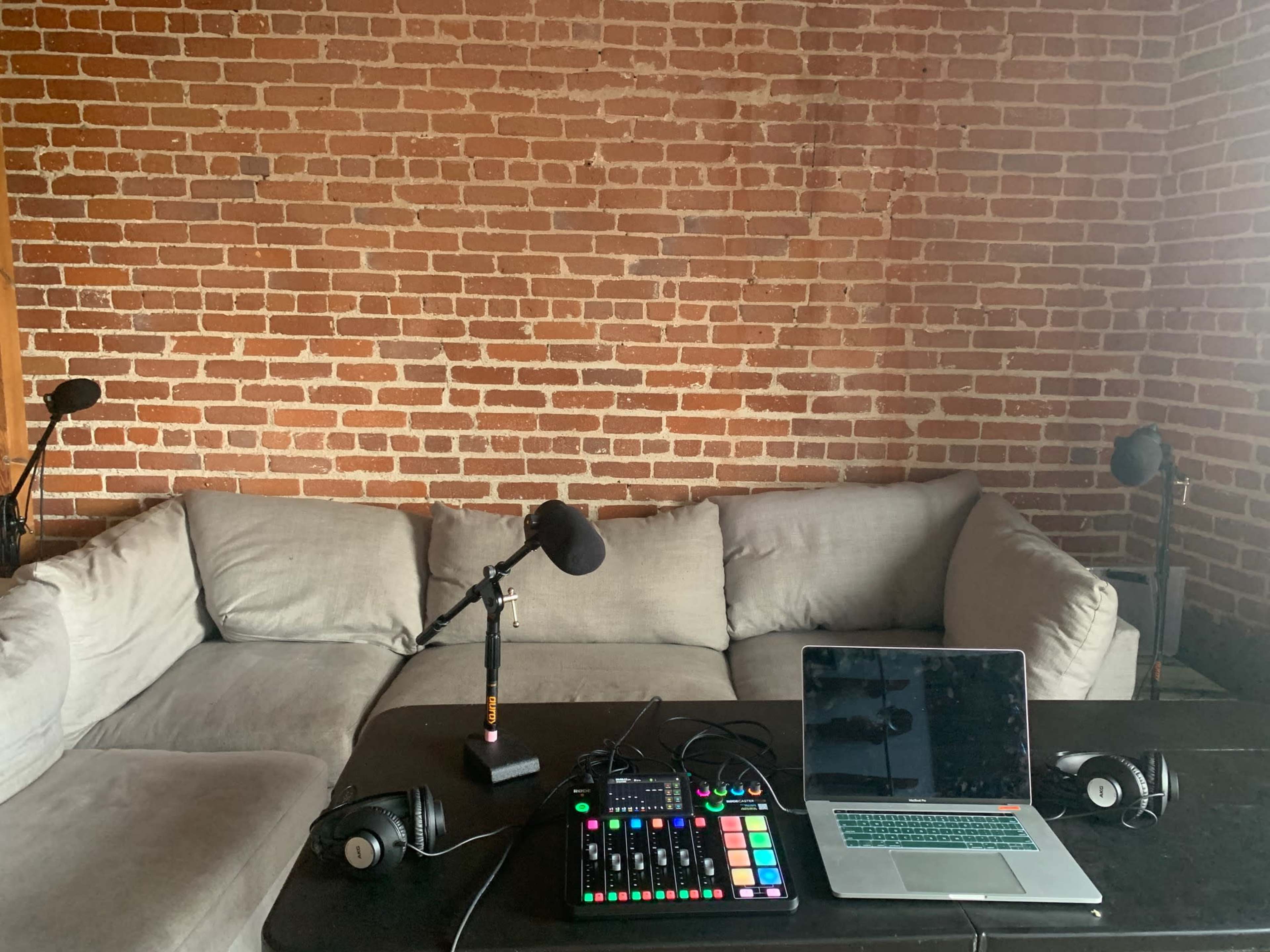 A gray sectional couch is positioned against a brick wall, with a laptop, audio mixer, headphones, and microphones set up on a black table in front.