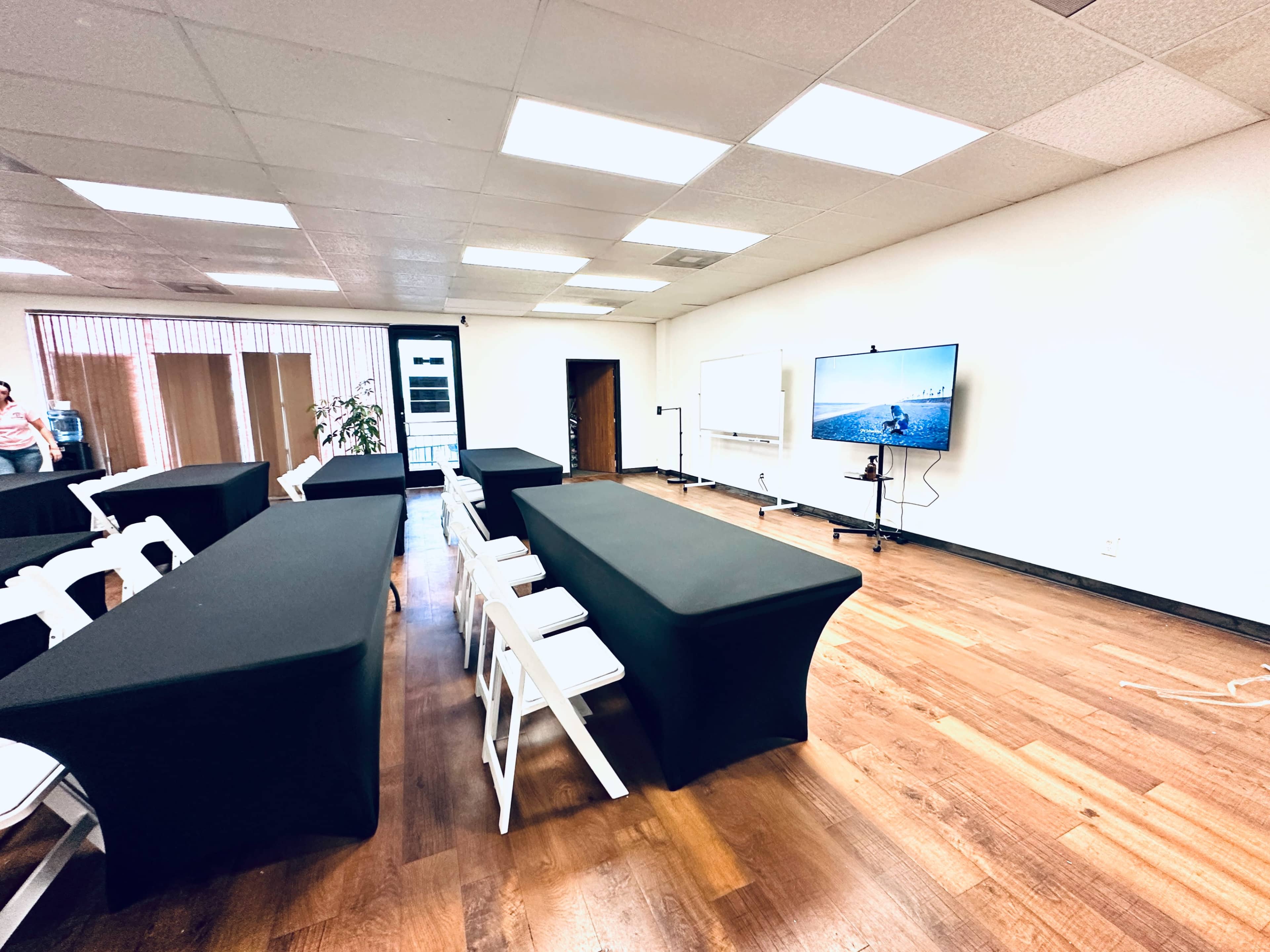 The image shows a meeting room with two rows of black-covered tables, chairs arranged neatly, and a large screen mounted on a wall.