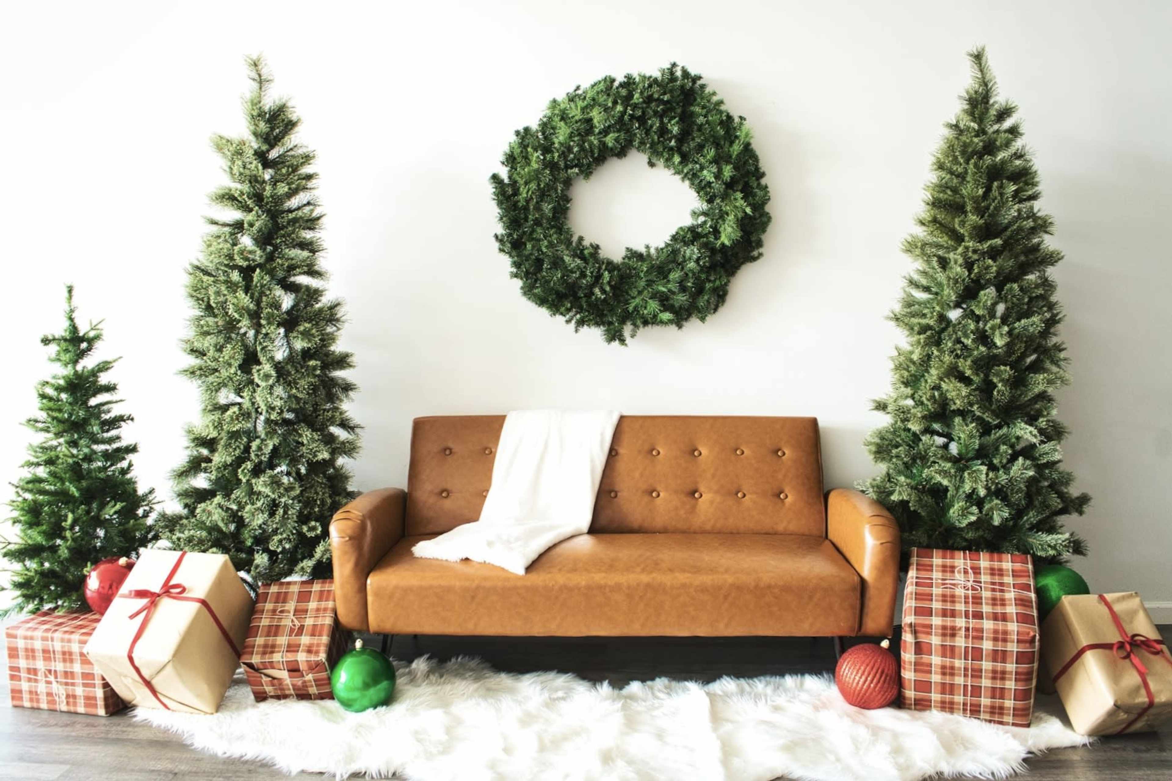 A brown leather sofa is positioned in front of two green Christmas trees and a large wreath, surrounded by wrapped gifts on a white fur rug.