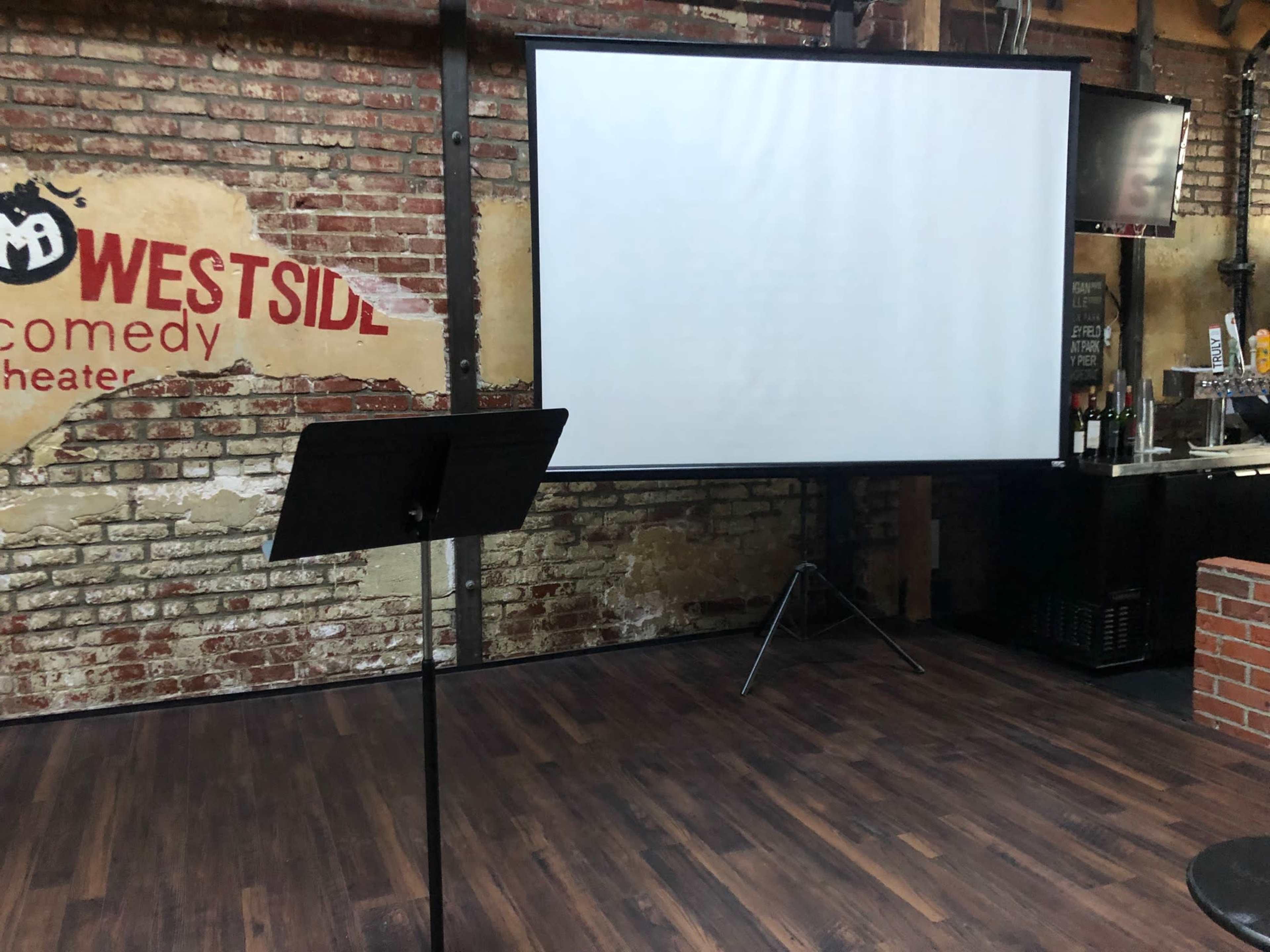 A black podium stands in front of a blank white projection screen in a brick-walled theater space.