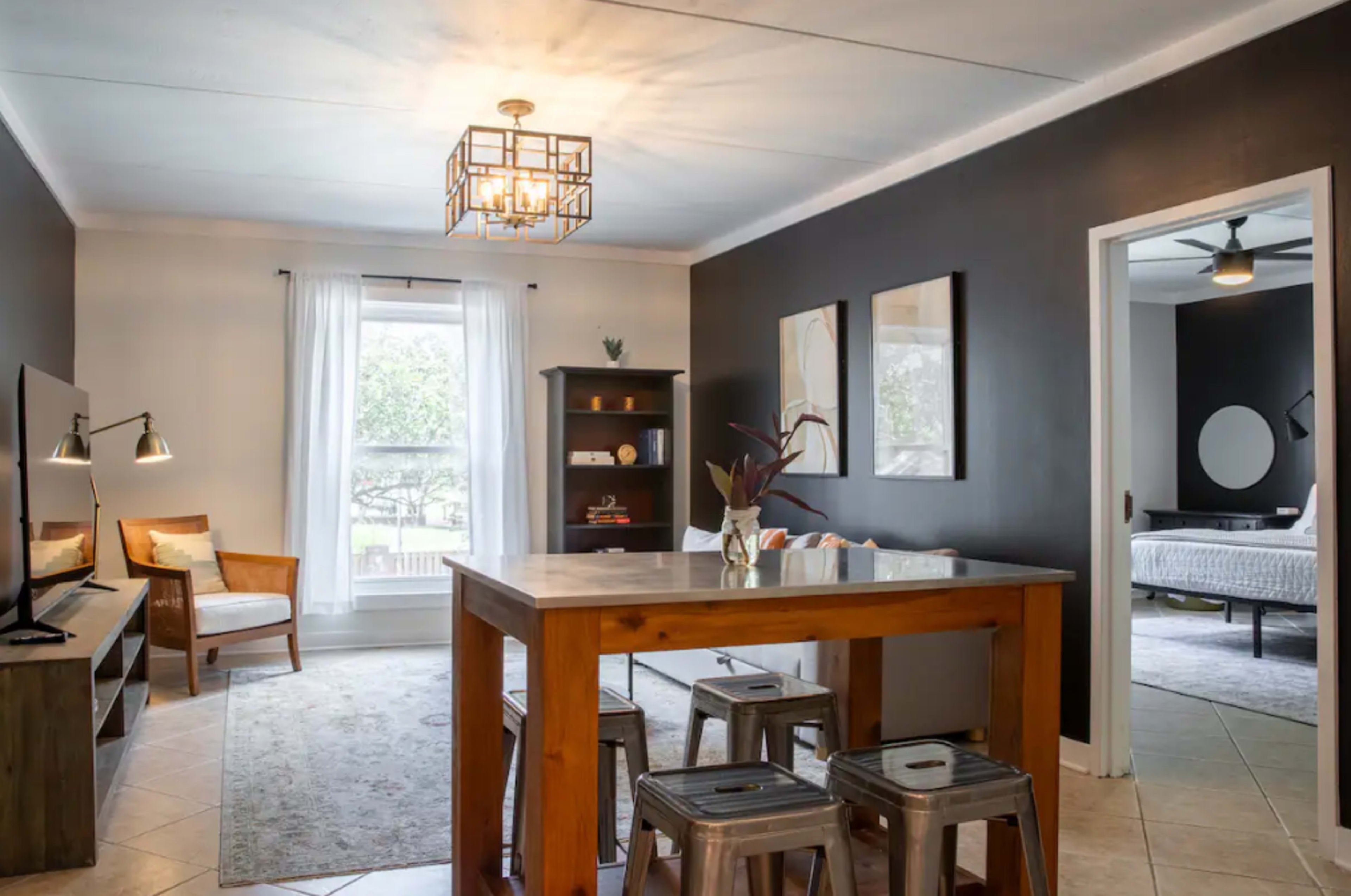 A living area with a wooden dining table, metal stools, a television, and a bookshelf, set against dark walls and featuring a large window with sheer curtains.