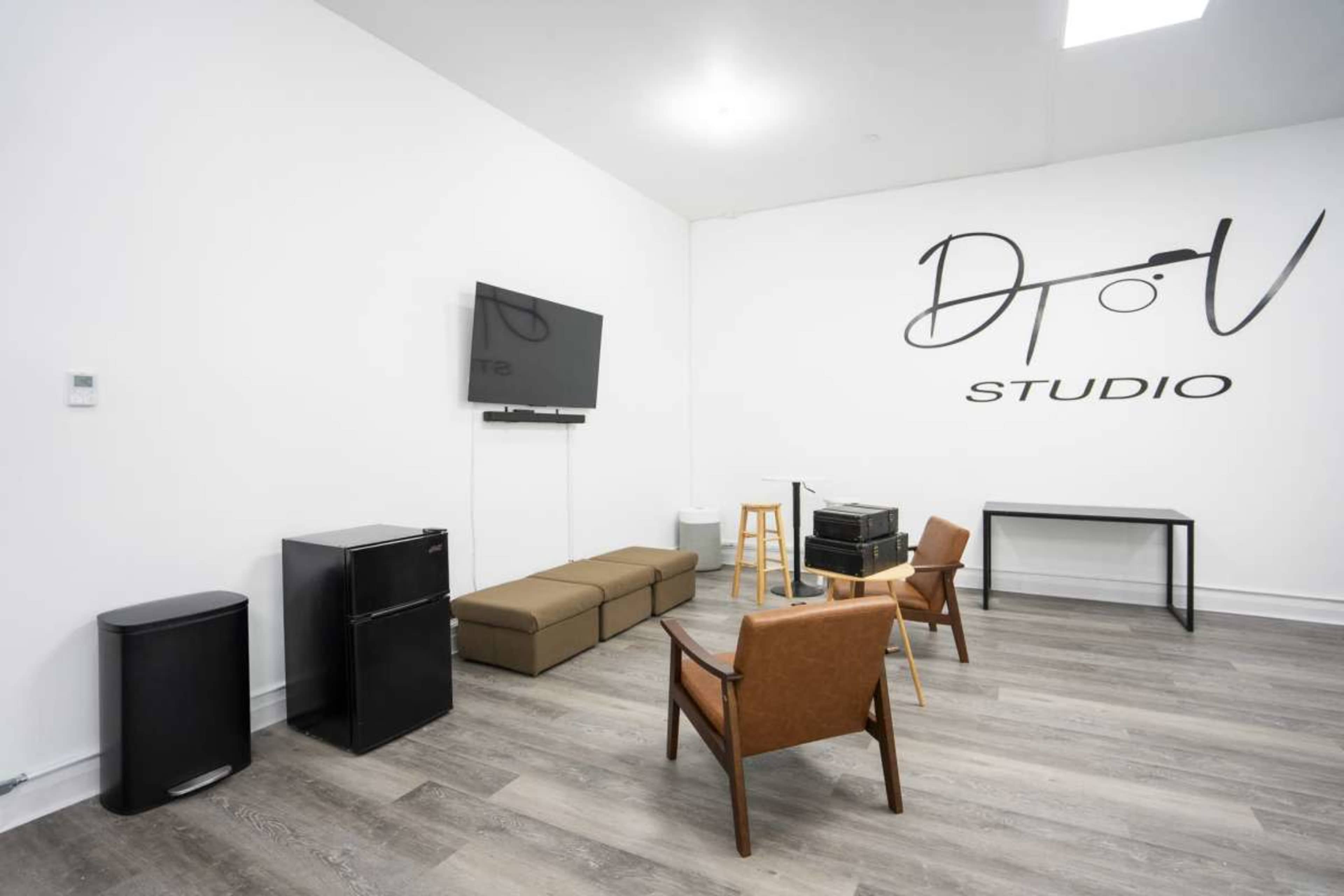 The image shows a minimalist studio space with a sofa, two chairs, a table, and a television mounted on the wall.