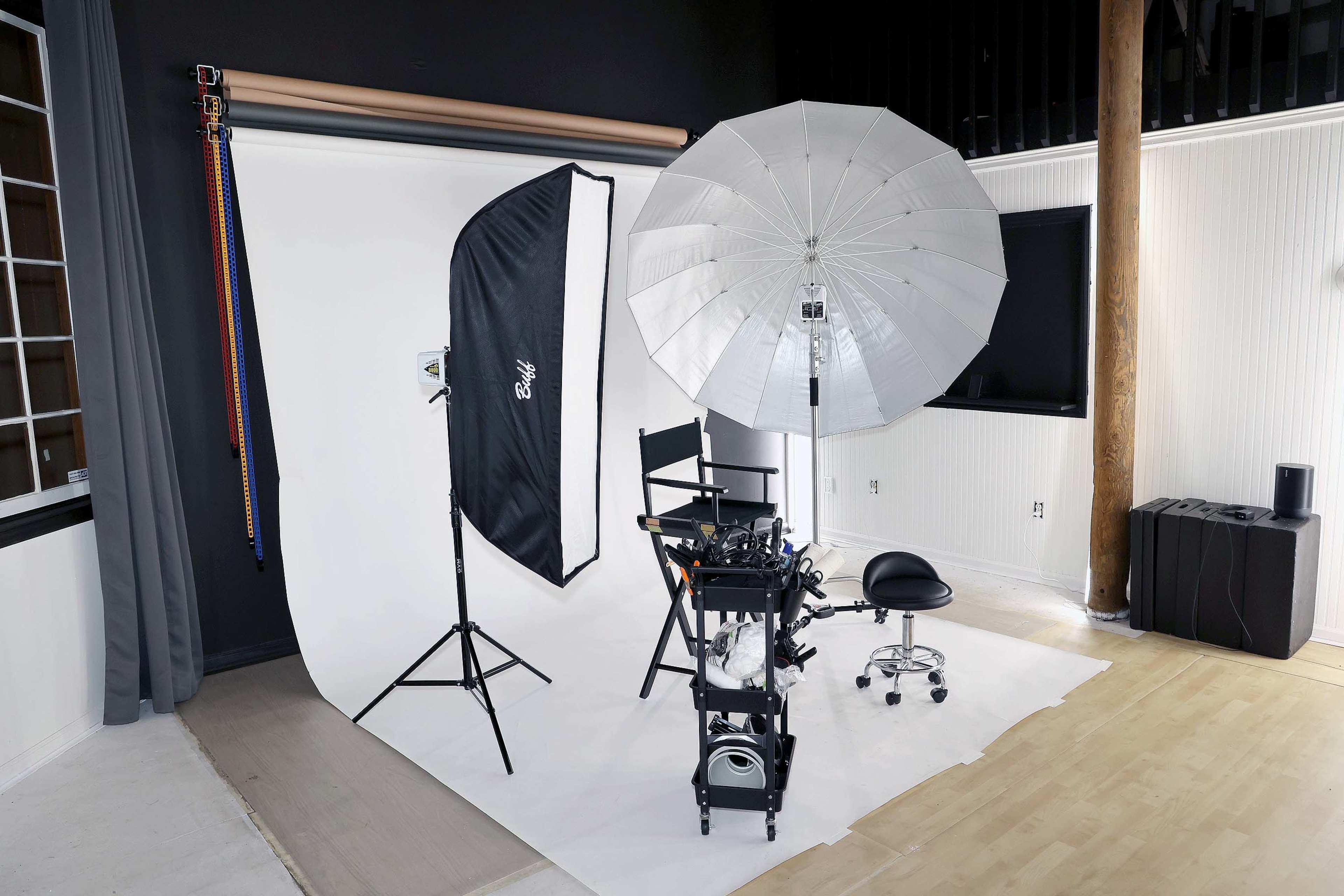 The image shows a photography studio setup featuring a white backdrop, lighting equipment including an umbrella and softbox, a director's chair, and various tools on a cart.