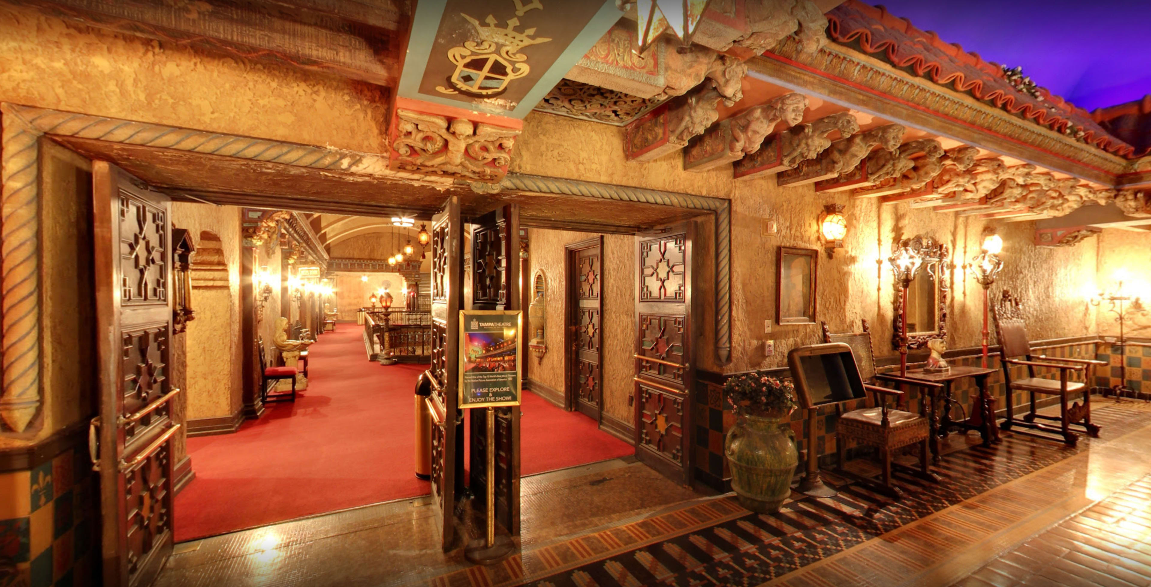 The image shows an ornate interior of a building with decorative walls, illuminated by vintage light fixtures and featuring a red carpet leading into a hallway.