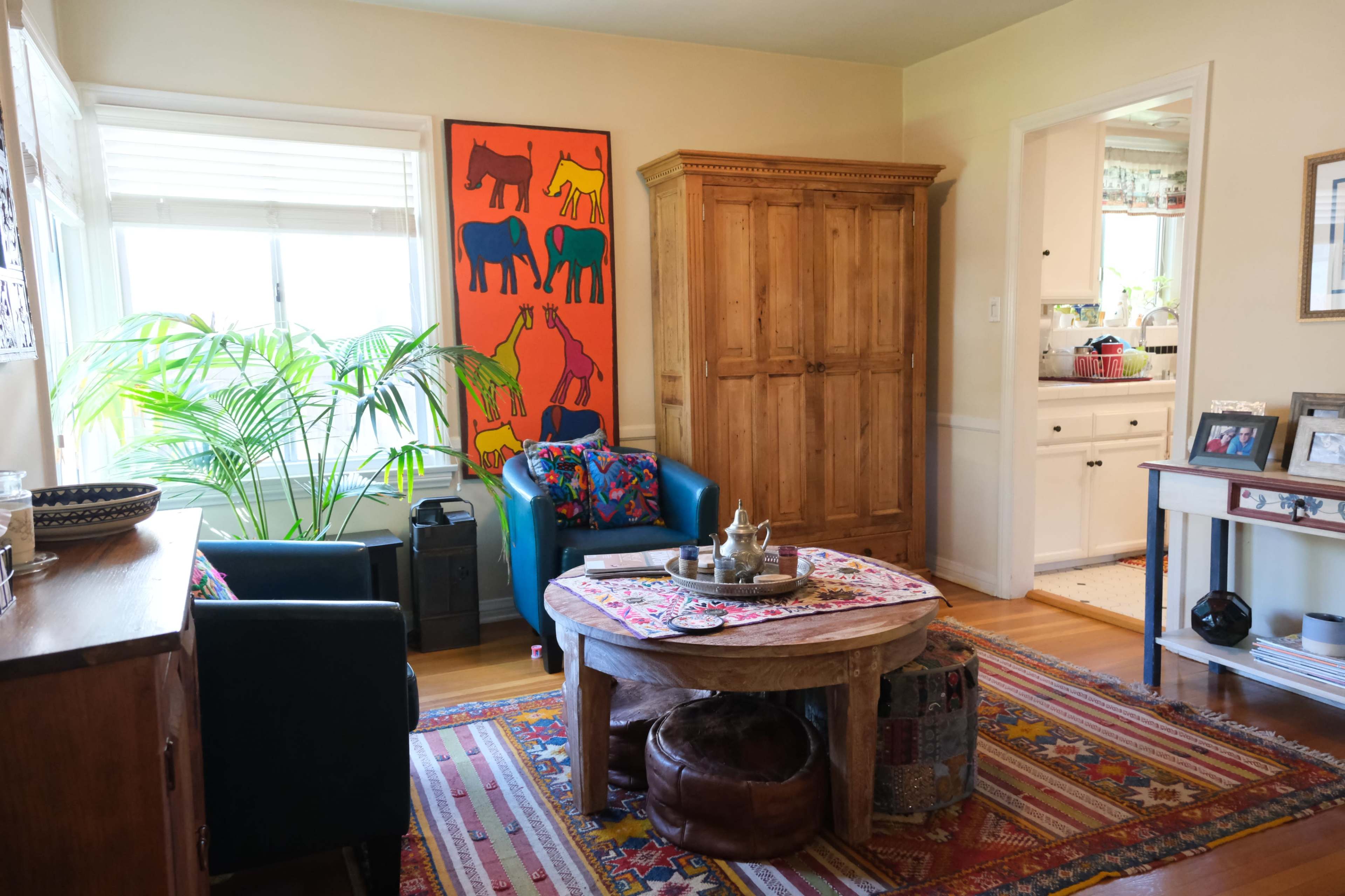 The room features a wooden coffee table surrounded by blue armchairs, a colorful area rug, and a large wooden cabinet against a wall with vibrant artwork.