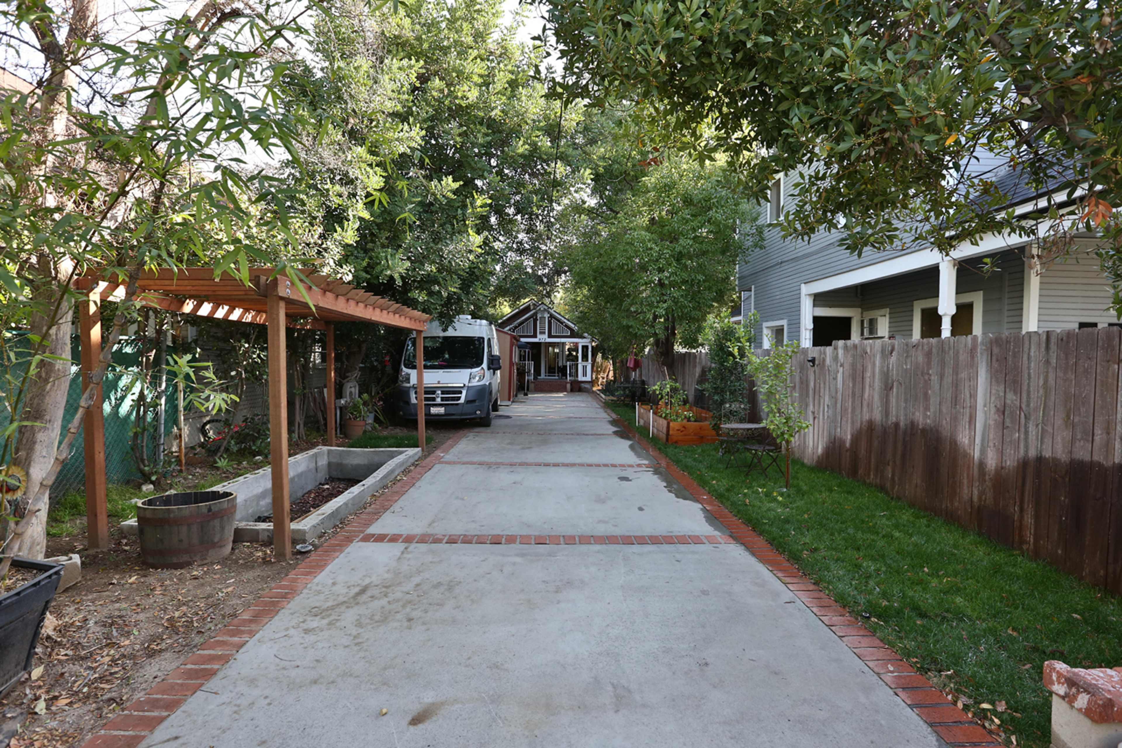 A paved driveway lined with trees leads to a building at the end, flanked by a wooden trellis and fenced areas.