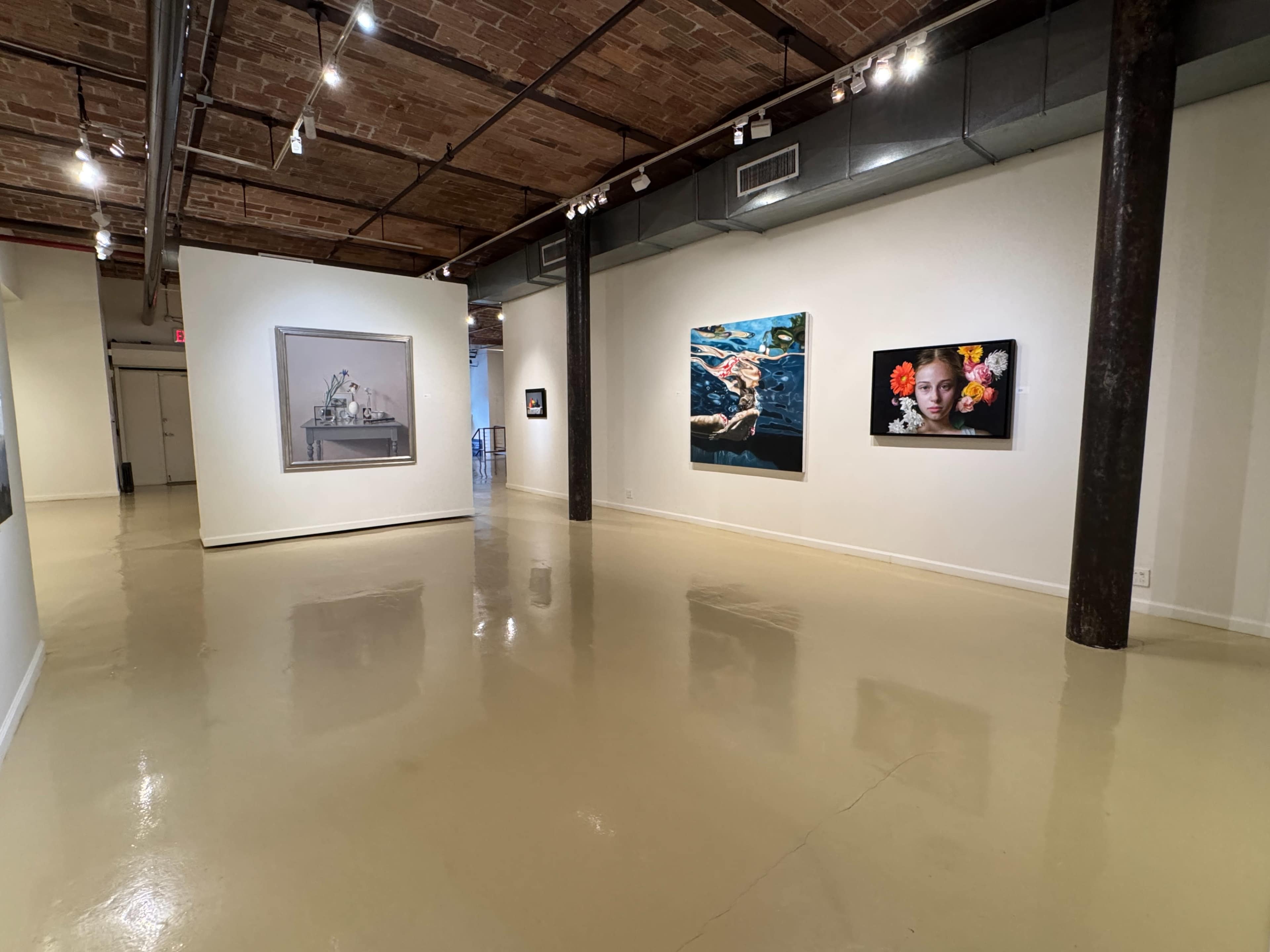 The image shows an art gallery with several framed artworks displayed on white walls and a polished floor under bright lighting.