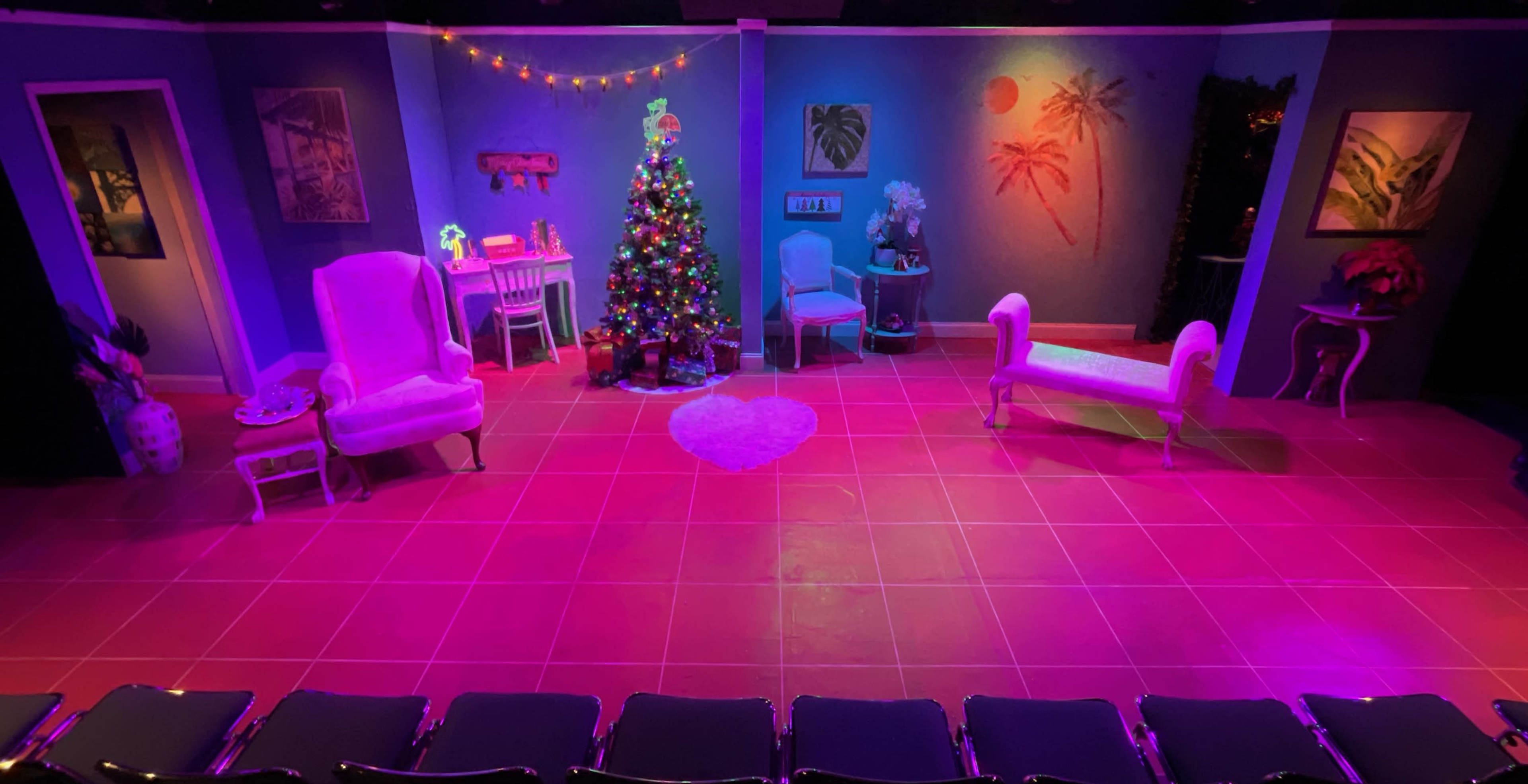 The image shows a brightly lit stage set decorated for a performance, featuring a Christmas tree, colorful artwork, and various seating arrangements.