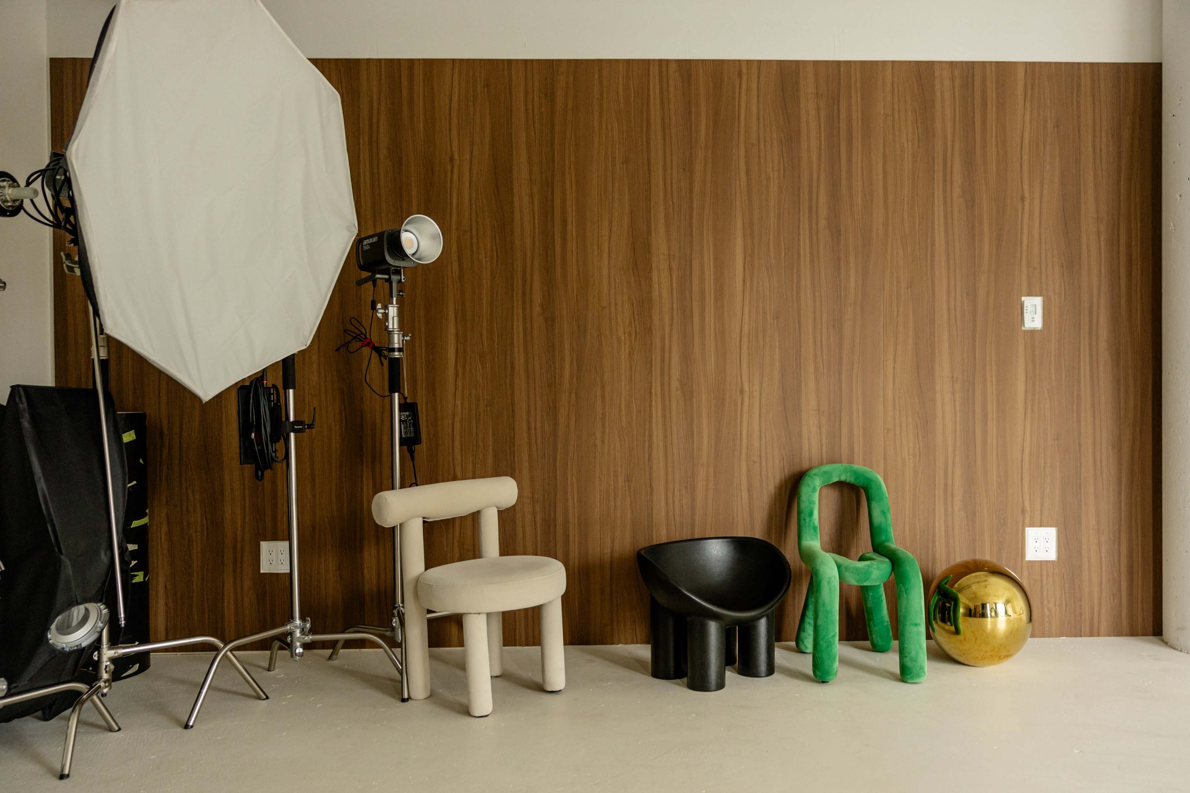 The image shows a studio setup with a large softbox light, a spotlight, and various modern chairs and a reflective ball against a wooden wall.