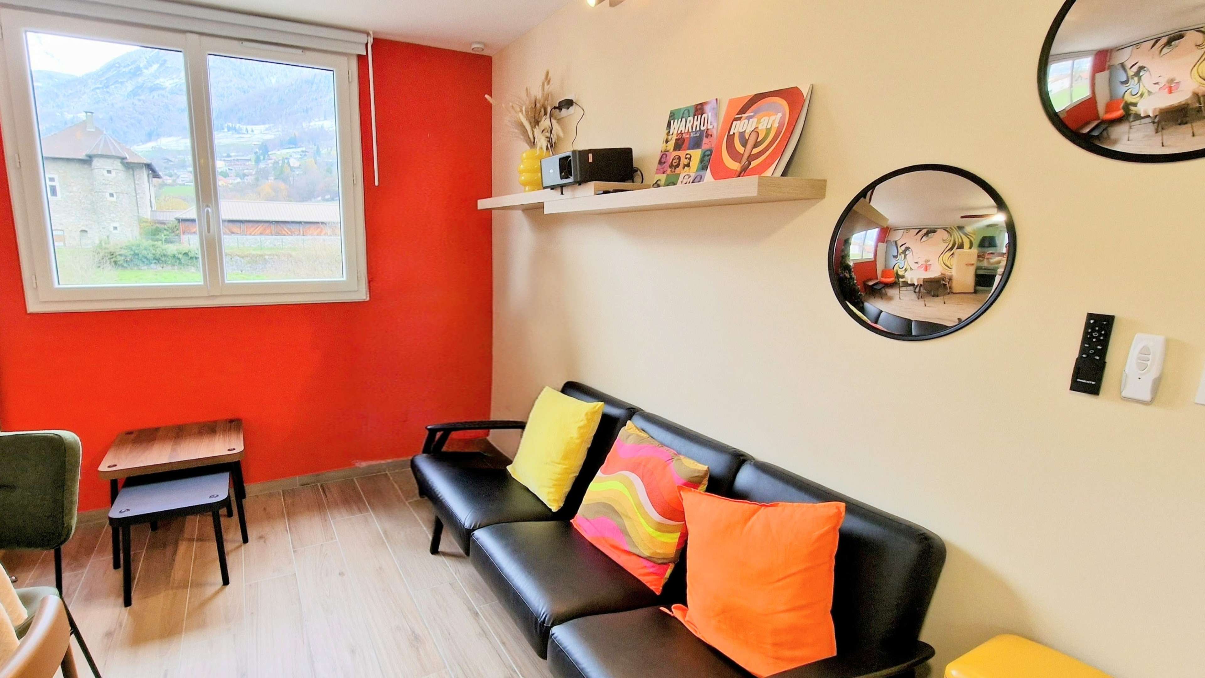 The image shows a cozy living room with a black couch adorned with colorful cushions, a wooden side table, and a window overlooking a mountainous landscape.