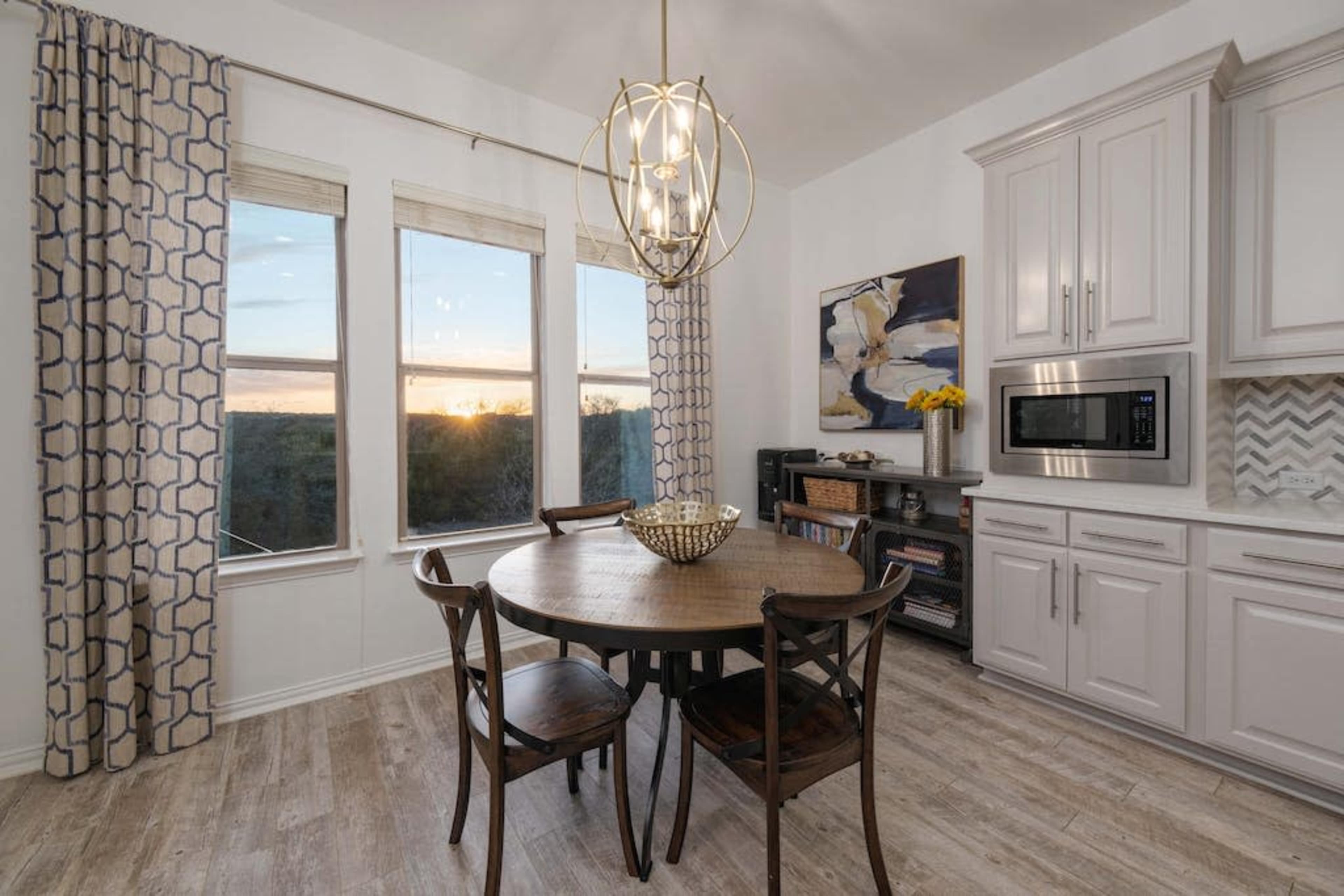 A round wooden table with four chairs is set near large windows, illuminated by a decorative light fixture, in a kitchen with light-colored cabinets and a patterned backsplash.